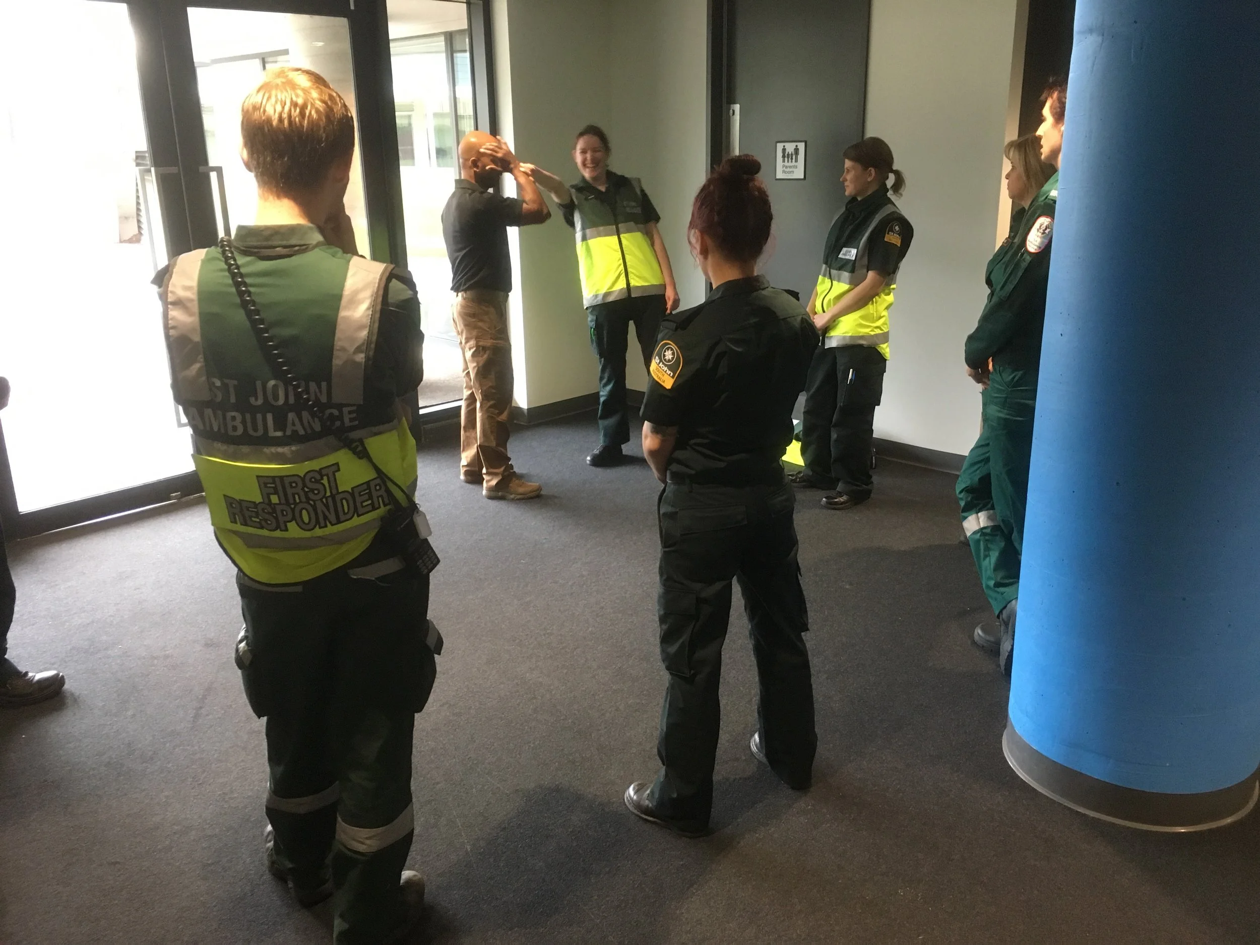 Group of emergency responders, including paramedics and medical personnel, standing inside a building near the entrance, some engaged in conversation or reaction, with a glass door and a sign indicating a children’s restroom in the background.
