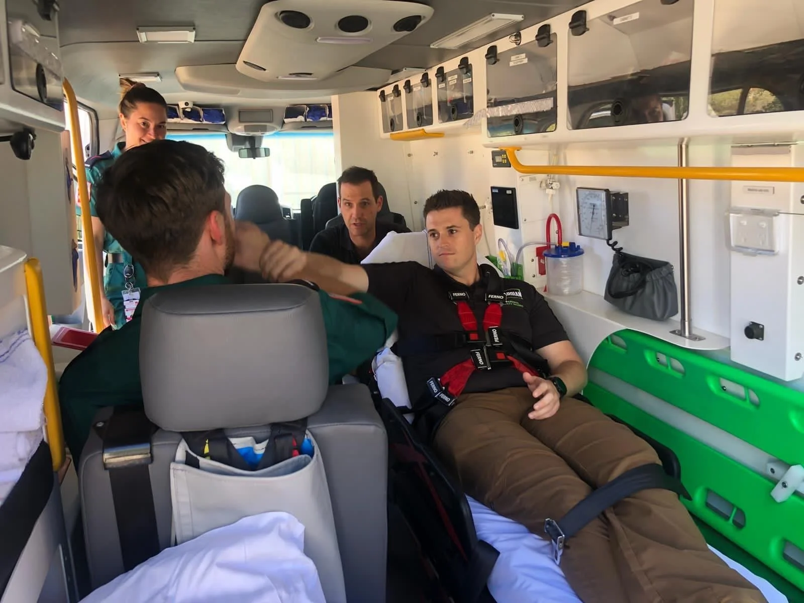 Inside an ambulance, a paramedic is lying on a stretcher receiving care from a staff member, while other medical personnel are present, preparing to help a patient.
