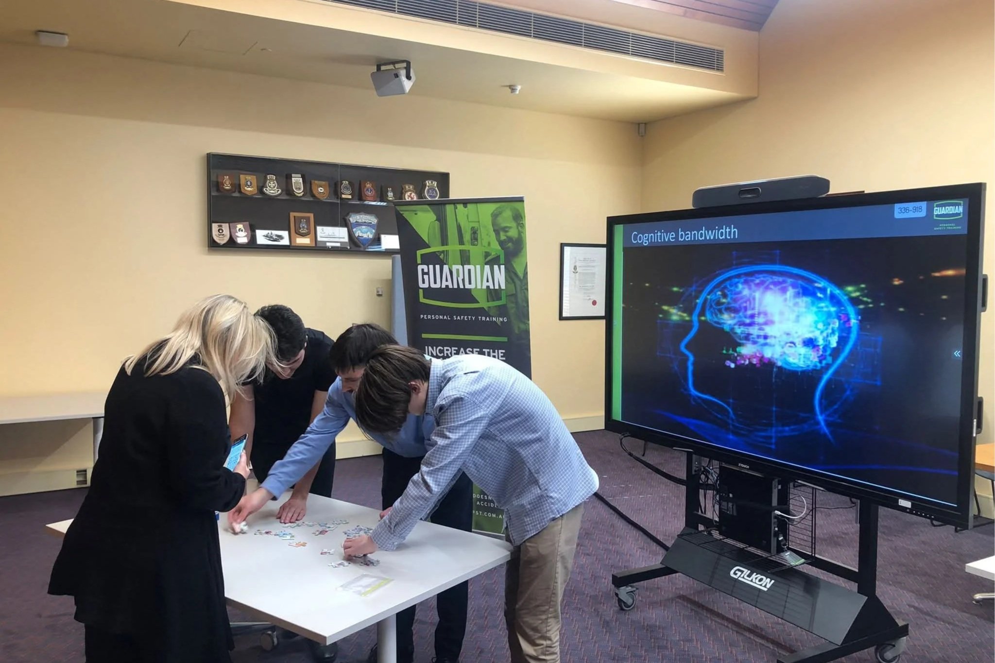 A group of people working together at a table in a conference room, with a large screen displaying "Cognitive bandwidth" and a digital brain graphic, and a banner for Guardian personal safety training in the background.