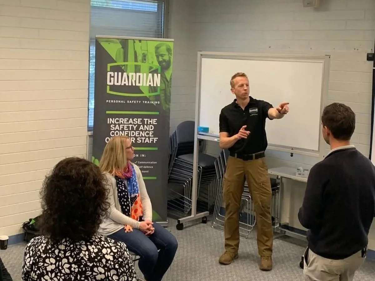 A man giving a safety training presentation to three people in a classroom, with a Guardian personal safety training banner in the background.