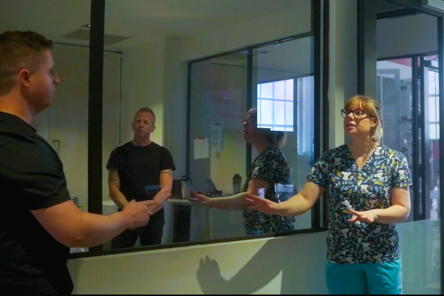 Four people having a discussion behind a glass partition in an office setting. Three women and one man. One woman has glasses and is speaking, another woman has crossed arms, and the man is holding a tablet.