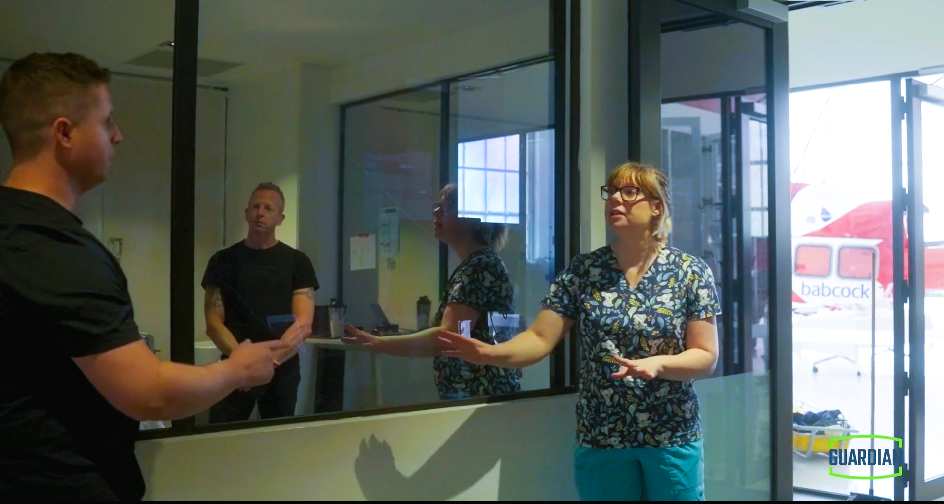 Four people, two women and two men, engaged in a conversation inside a building near a glass window and door. One woman is gesturing with her hand while speaking to an individual outside. The background shows a red bus outside. A