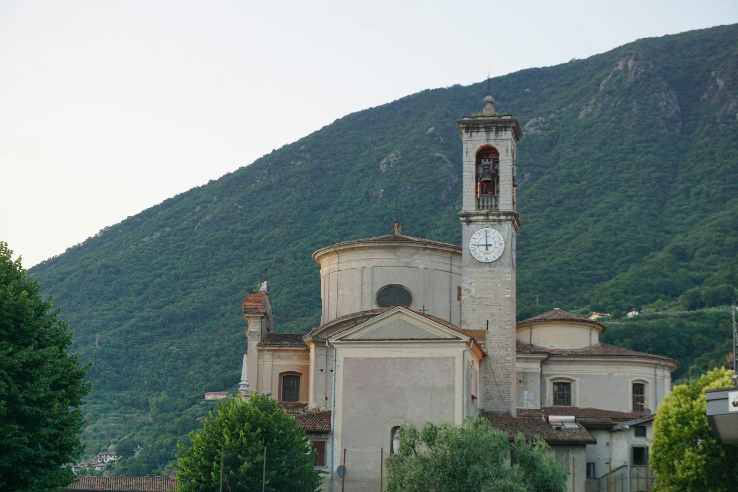 Una chiesa con un alto campanile e un orologio, circondata da alberi verdi e con montagne sullo sfondo.