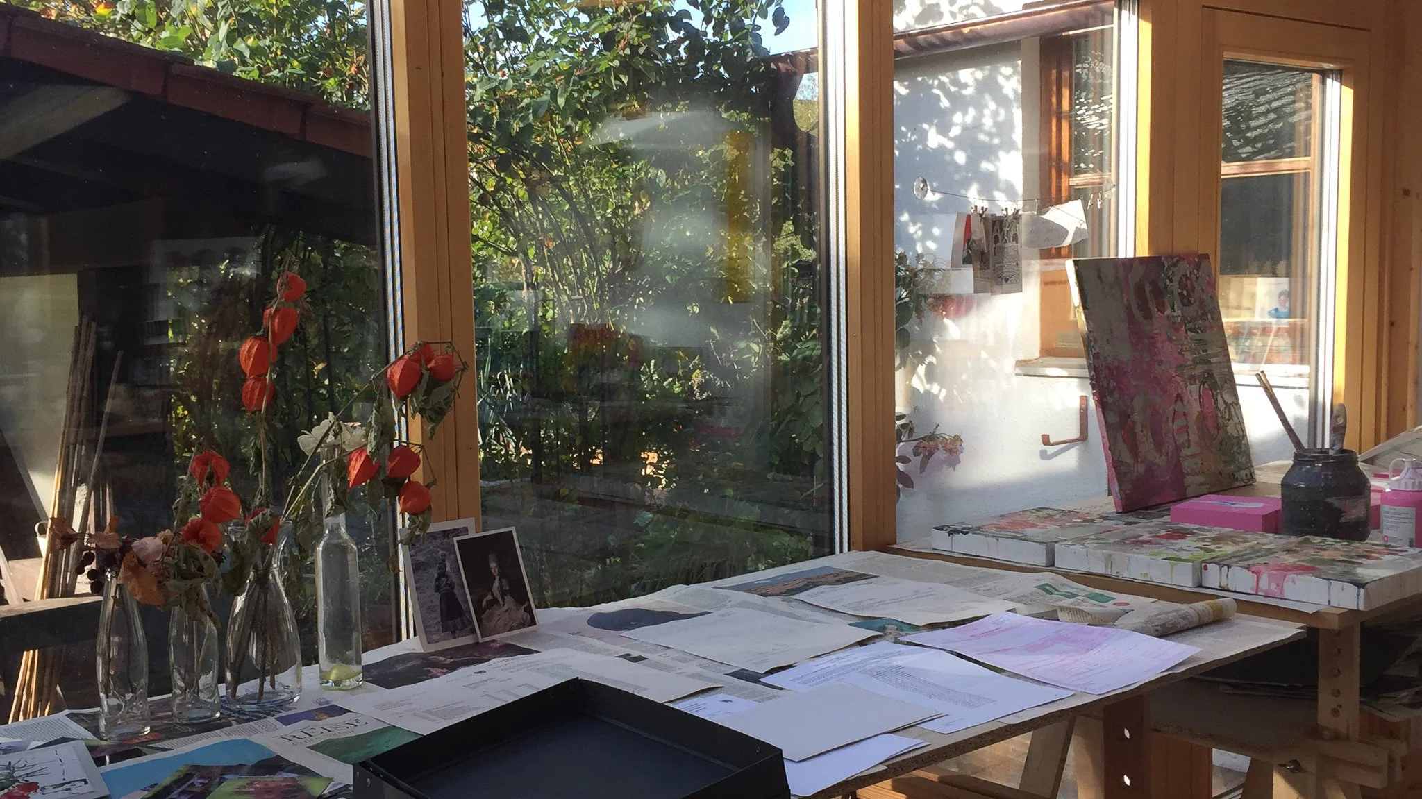 Ein Tisch mit geöffneter Zeitschrift, Fotos, Gläser mit roten Blumen, und mehreren Papieren, vor einer großen Fensterfront mit Blick auf einen Garten, im Sonnenlicht.