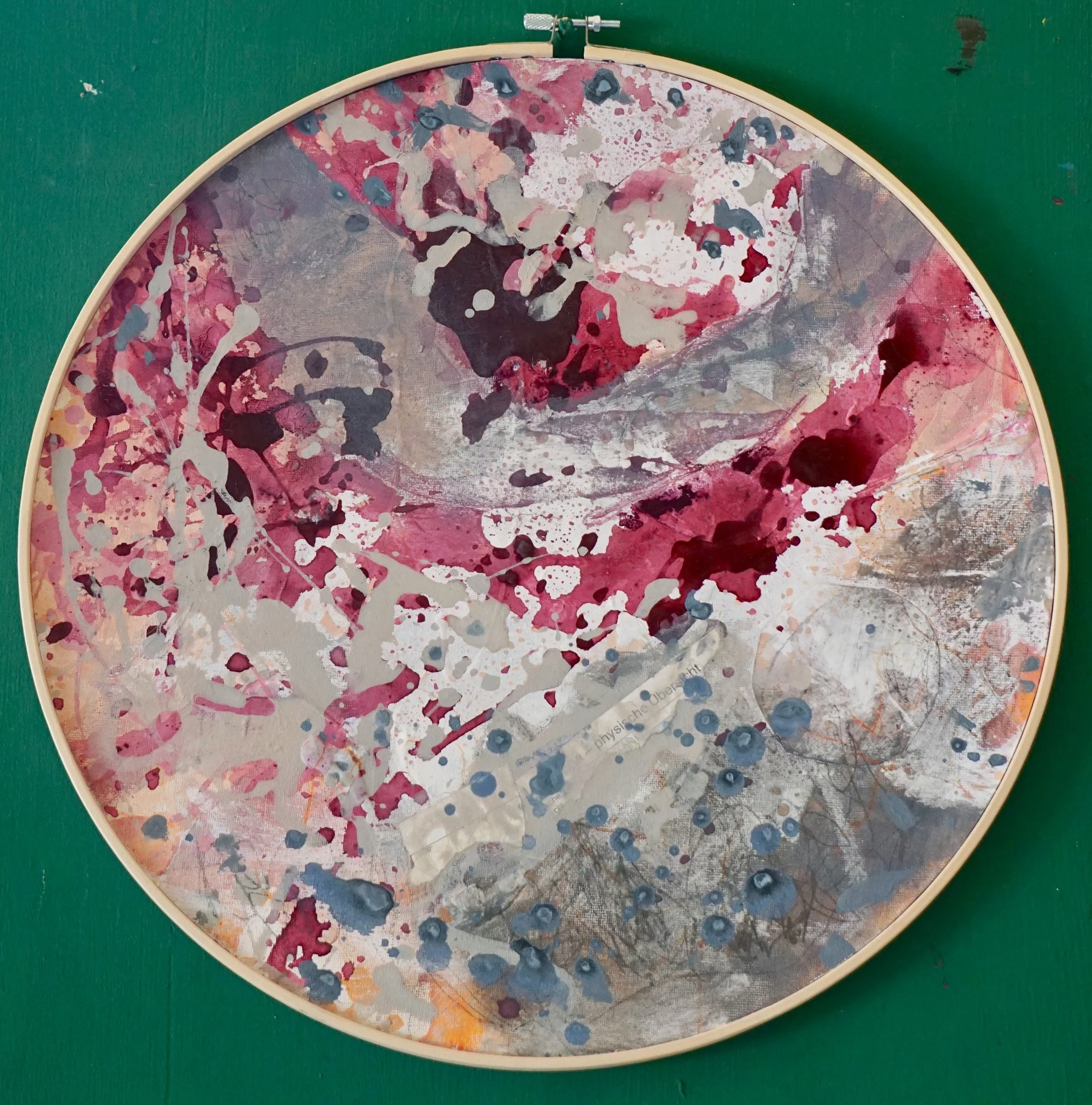 Abstraktes, farbenfrohes Kunstwerk in einem runden Rahmen, mit pinken, grauen, schwarzen und blauen Farbspritzern und Mustern auf einer grünen Wand.