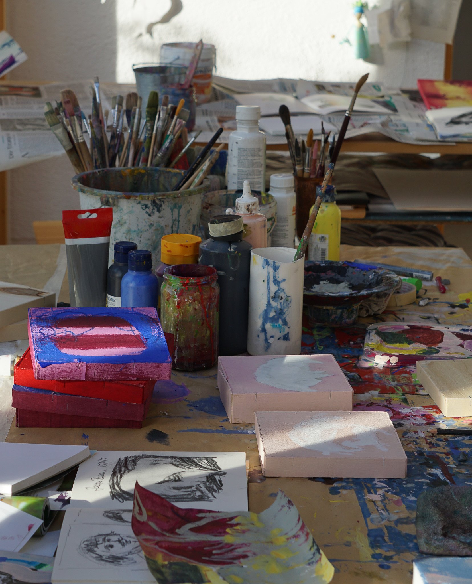 Ein Tisch mit verschiedenen Malutensilien wie Farben, Pinsel, Malpaletten und Skizzen, umgeben von Papieren und Kunstwerken.