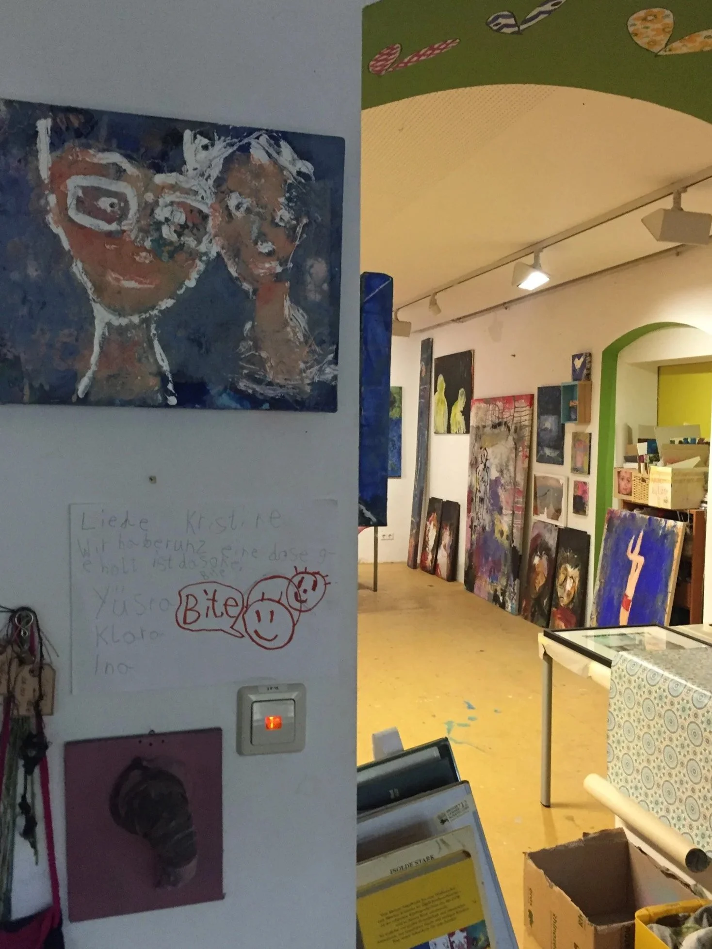 Blick in eine Kunstgalerie mit mehreren Gemälden an der Wand, einem Bücherregal und einem Tisch.