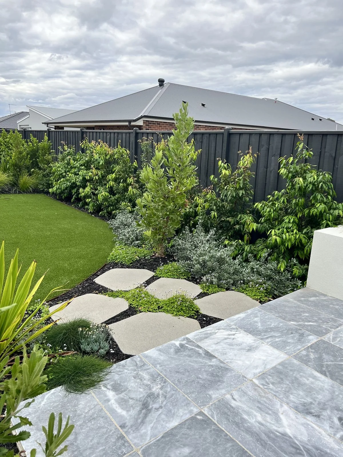 A landscaped backyard with a slate patio, a green lawn, a variety of shrubs and small plants, stepping stones, and a black wooden fence, under a cloudy sky.