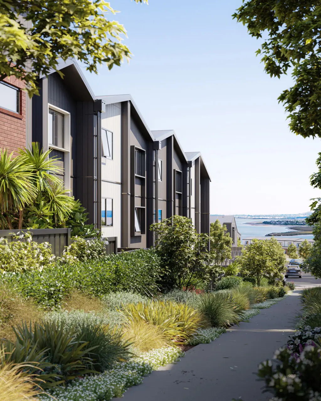 View of modern residential buildings with dark and white siding, surrounded by lush greenery and a well-maintained sidewalk, overlooking a body of water in the distance on a clear, sunny day.