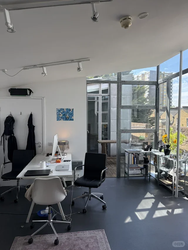 Modern office space with a white desk, two black chairs, and a white chair, near large windows with a city view and a small balcony with plants.