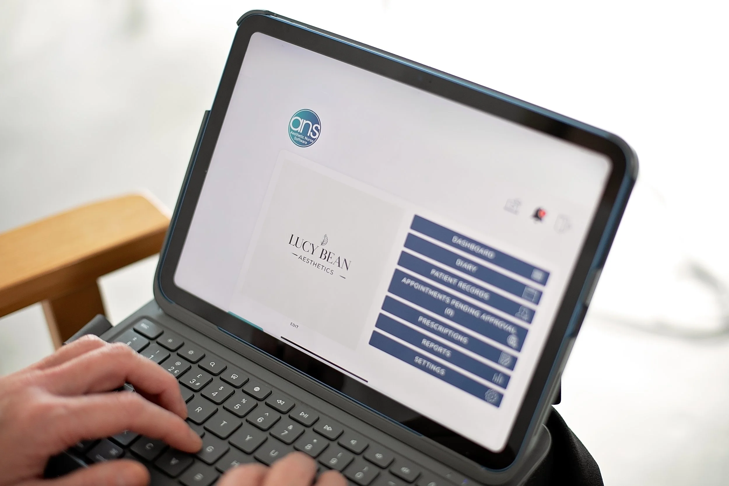 A person using a tablet with a keyboard displaying the Lucy Bean Aesthetics software interface, showing options like dashboard, diary, patient records, appointments, prescriptions, reports, and settings.