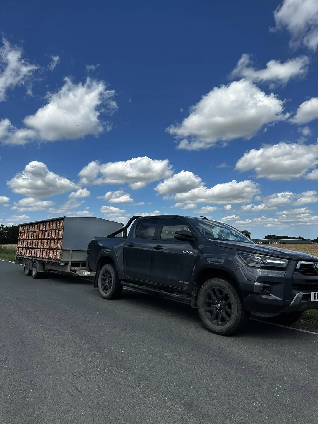 Black Toyota Hilux pickup truck pulling a trailer with a large metal container, parked on the side of a rural road under a blue sky with scattered clouds.