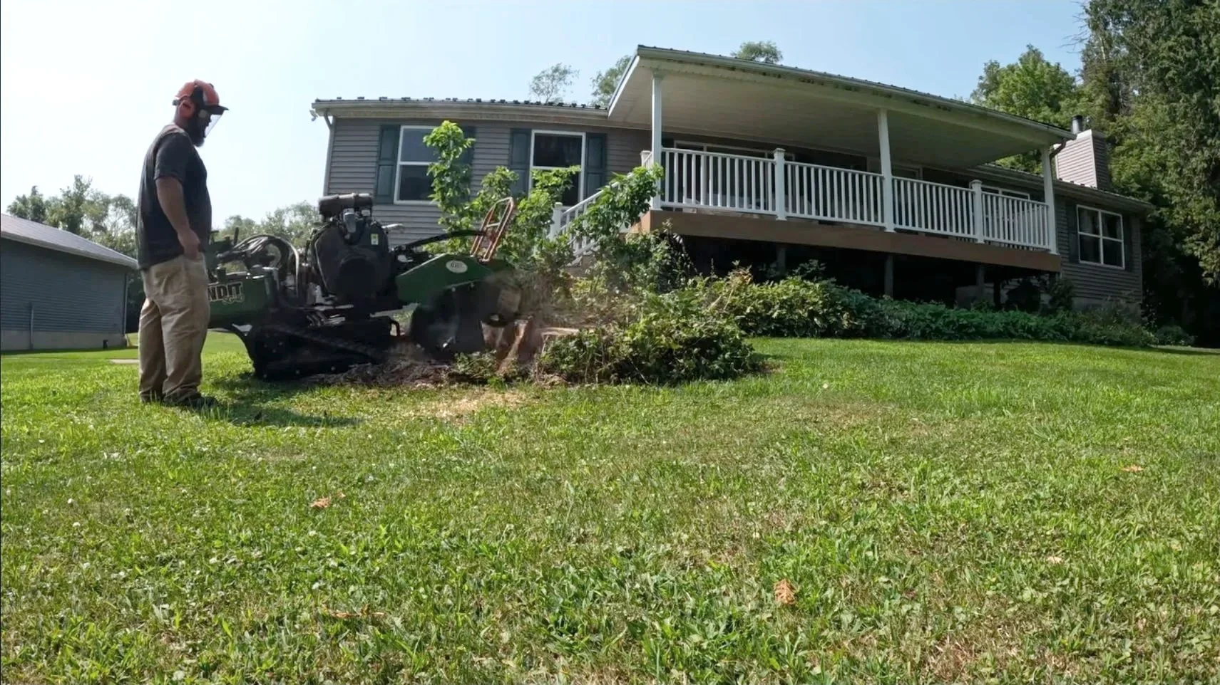 A man stands next to a stump grinder working on removing a tree stump from a yard, with a house and trees in the background.