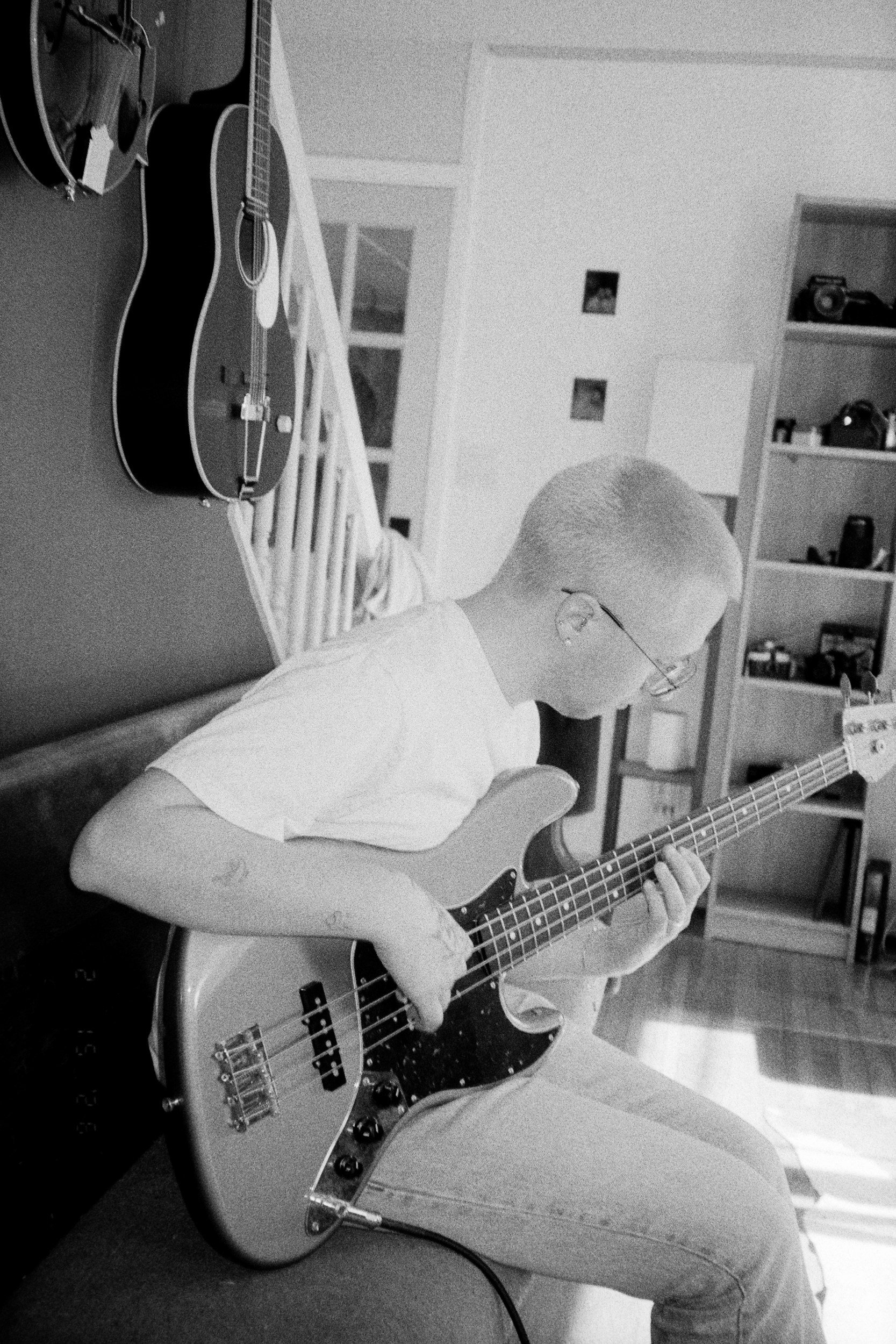 A person playing an electric bass guitar while sitting on the floor in a room, with a shelf and guitars on the wall in the background, in black and white.