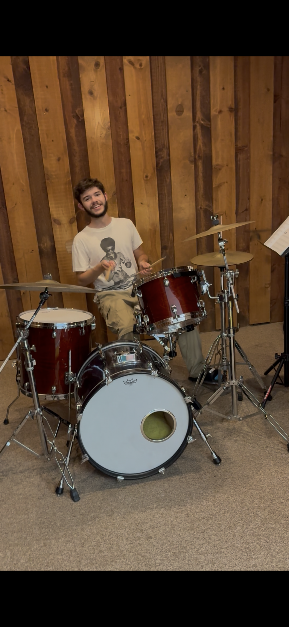 A young man smiling and sitting behind a red drum set in a room with wooden walls. He is holding drumsticks and appears to be ready to play.