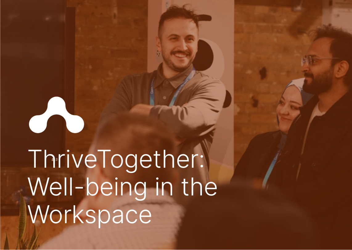 A group of people at a workspace or conference, with one man smiling and others listening. There is a brick wall in the background, and the people are wearing badges, suggesting a professional or networking event. ThriveTogether: Well-being in the Workspace