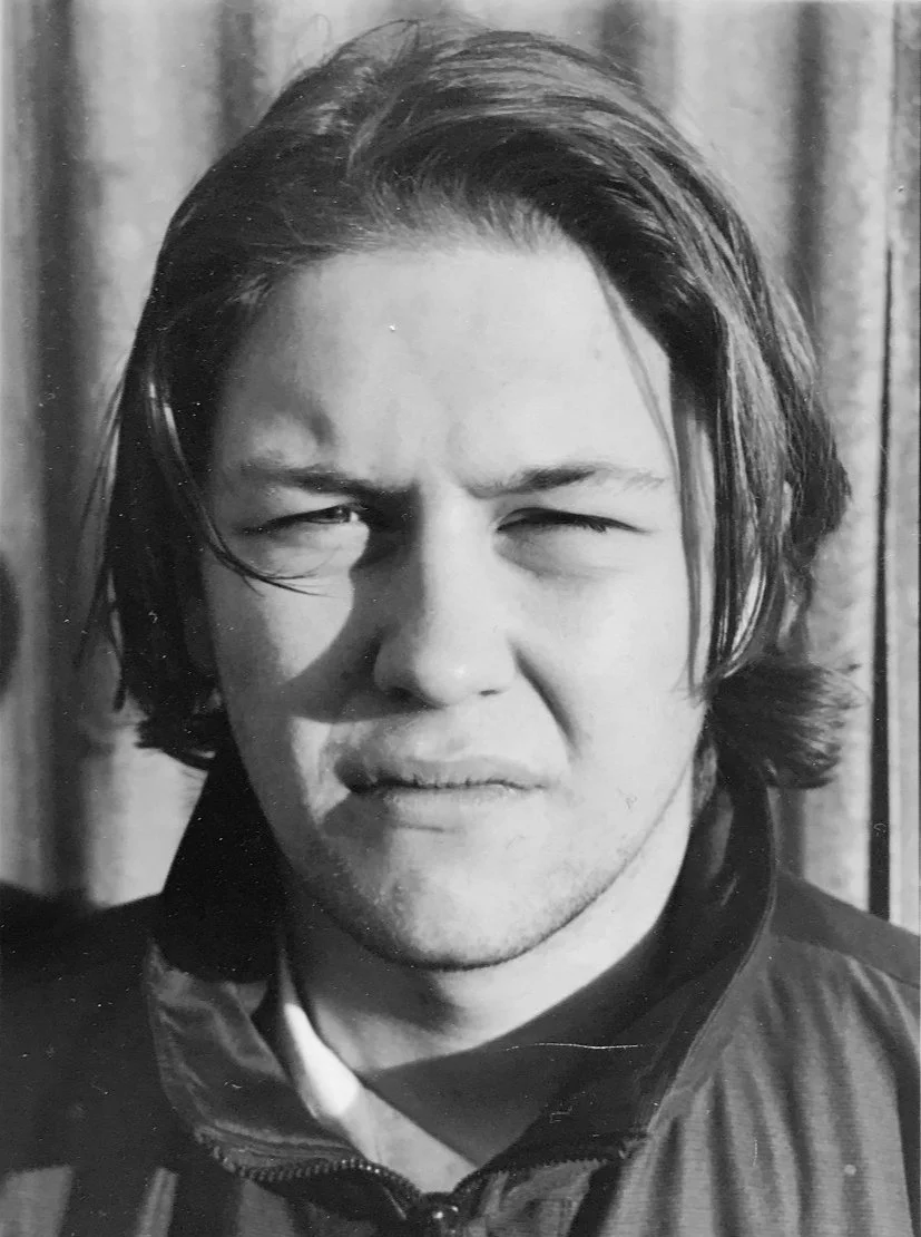 Black and white photo of a young man with shoulder-length hair, squinting with a serious expression, wearing a zip-up jacket.