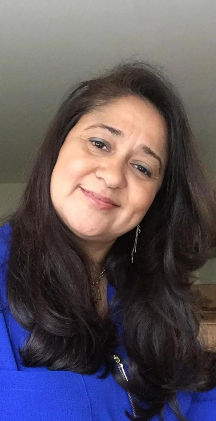 A woman with long dark hair, smiling, wearing a blue top and silver jewelry, indoors.