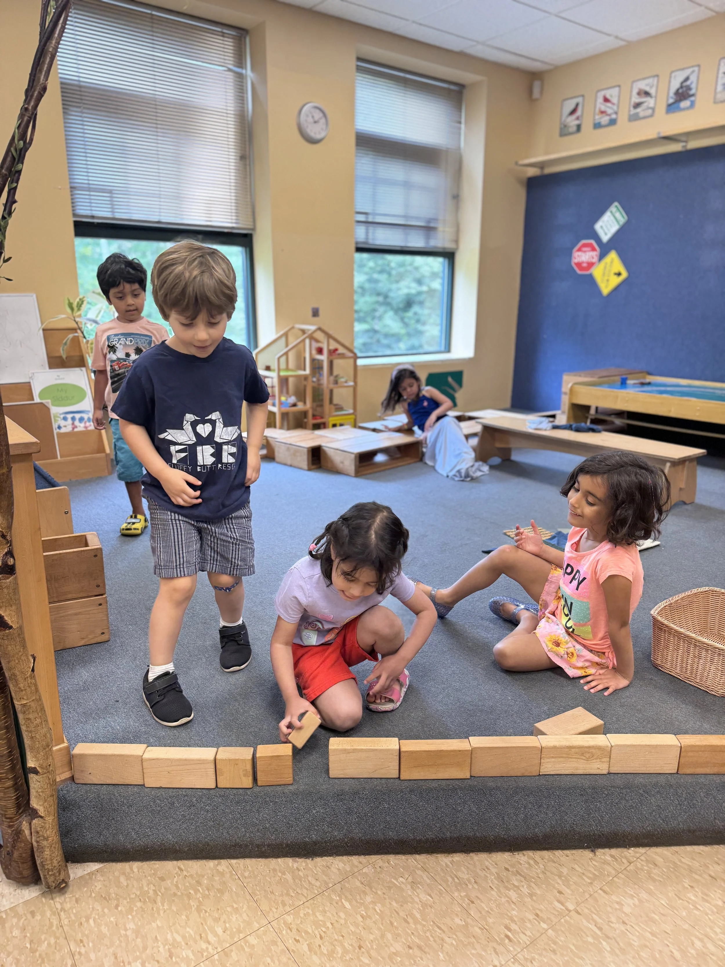 Child playing with wooden blocks on a school carpeted area inside a classroom with large windows, other children in the background, and educational posters on the wall.