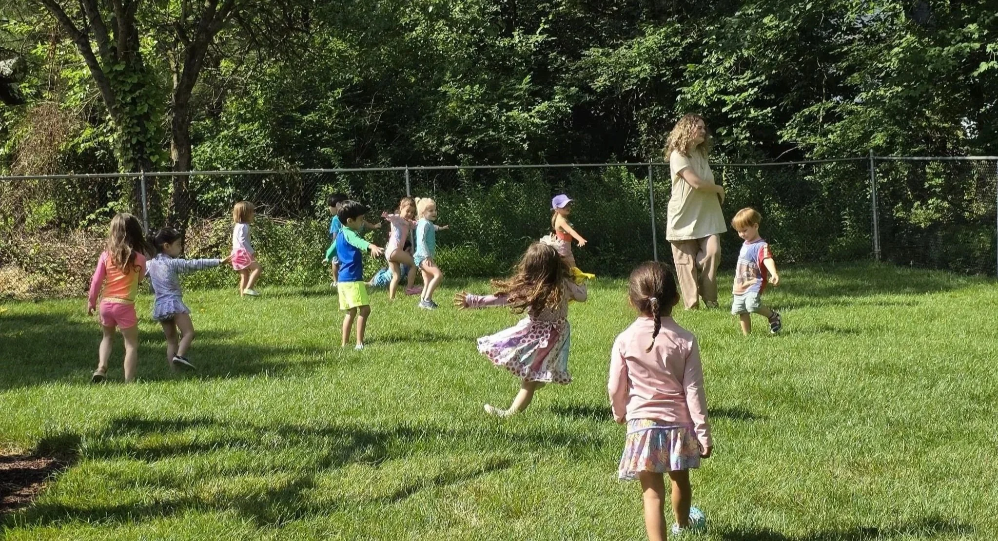 Children playing a game of tag or chase on a grassy field, supervised by an adult woman, with trees and a fence in the background.