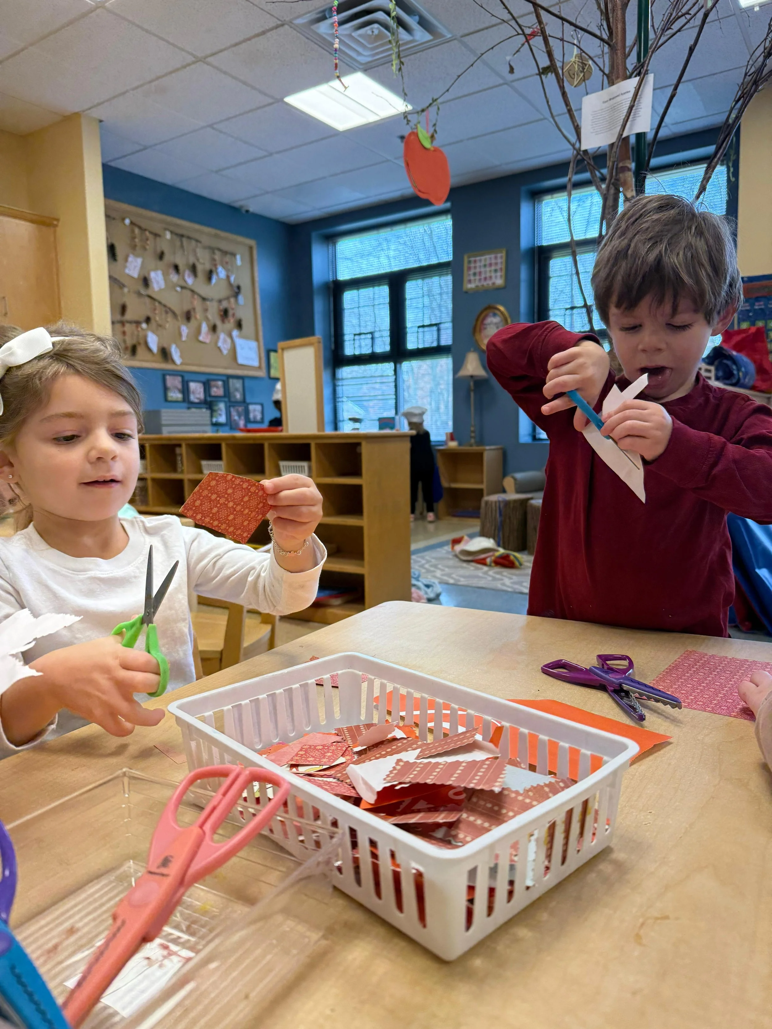 Two children, a girl and a boy, sitting at a table engaging in arts and crafts, cutting paper with scissors.