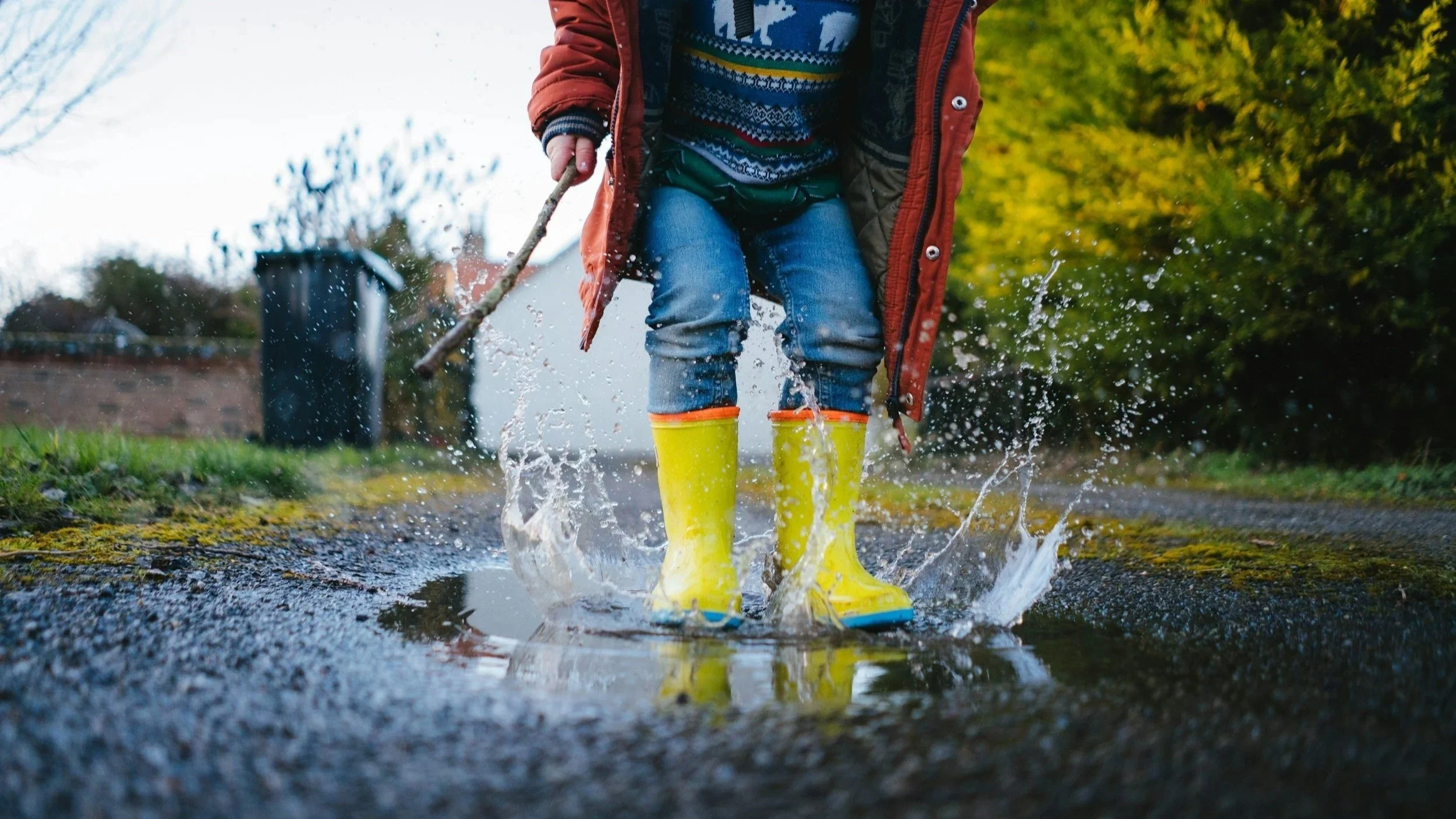 Child wearing yellow rain boots and jeans, splashing in a puddle on a rainy day, with a person holding a stick in the background.