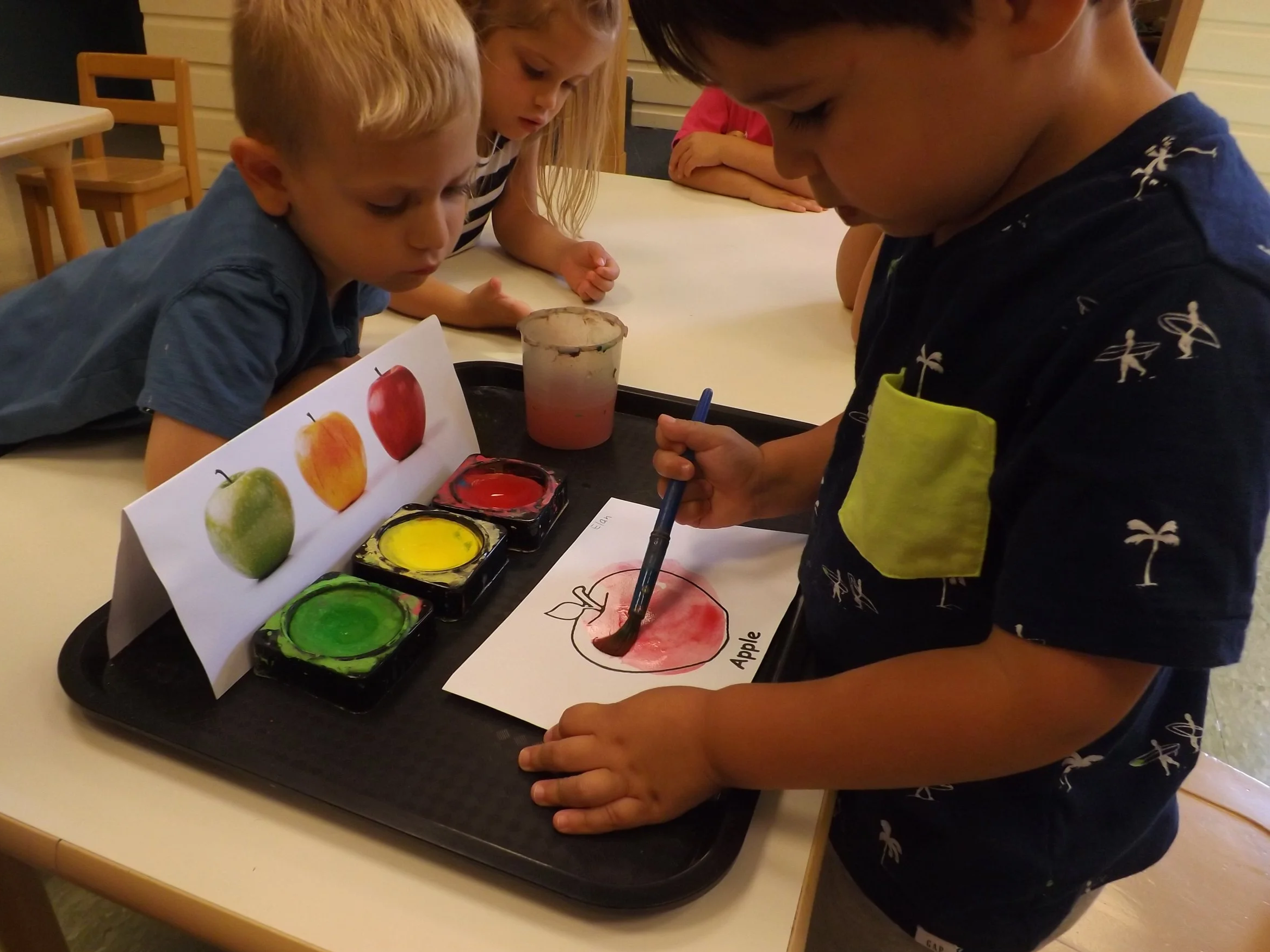 Children at a table during an art activity, painting and coloring pictures of apples with watercolors.