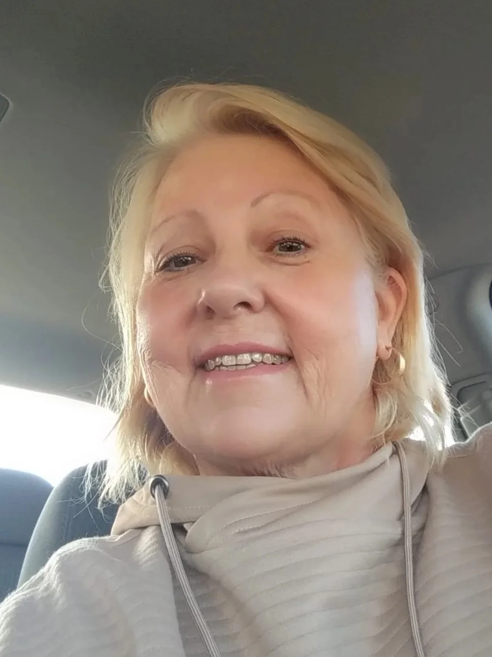 Close-up of a middle-aged woman with blonde hair, smiling inside a vehicle.