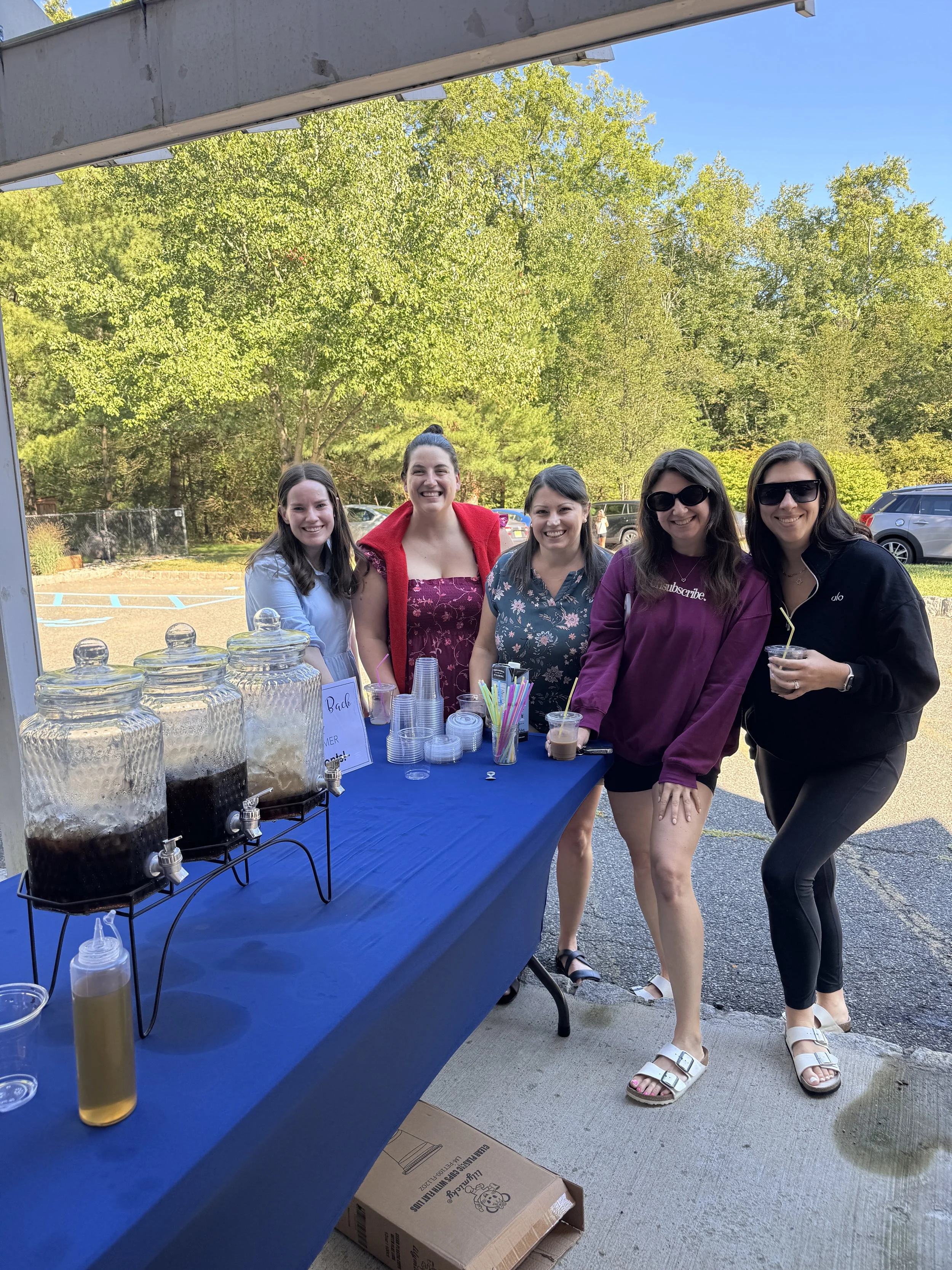 Five women stand outdoors in front of a table with drinks and cups, smiling for the camera during a sunny day. Green trees and parked cars are visible in the background.