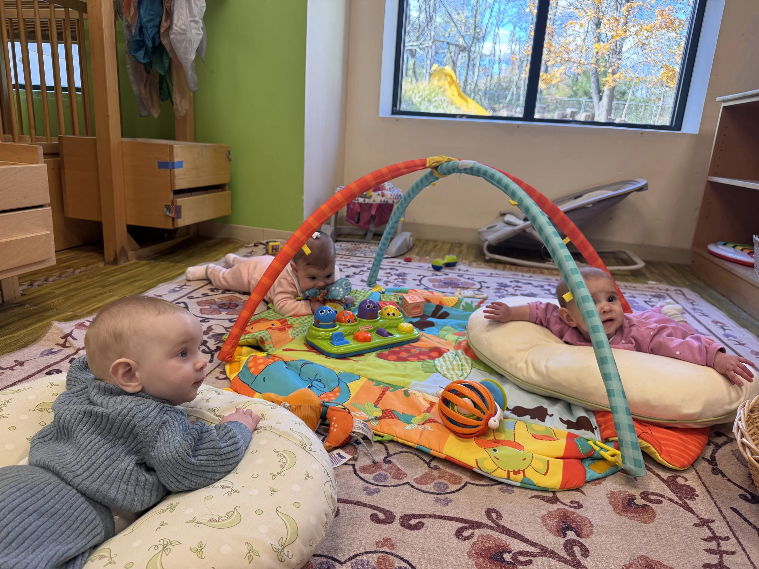 Three babies lying on the floor around a colorful play mat with toys, in a room with a window showing autumn trees outside.