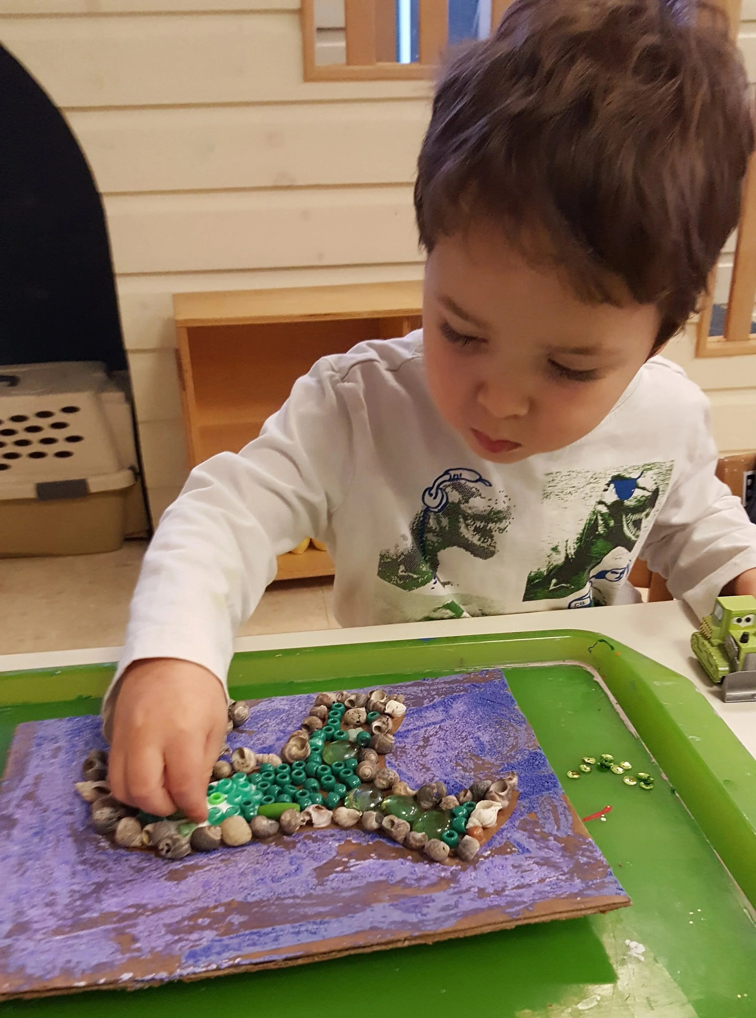 A young boy is working on a craft project with seashells, green beads, and a piece of cardboard painted purple, placed on a green tray.