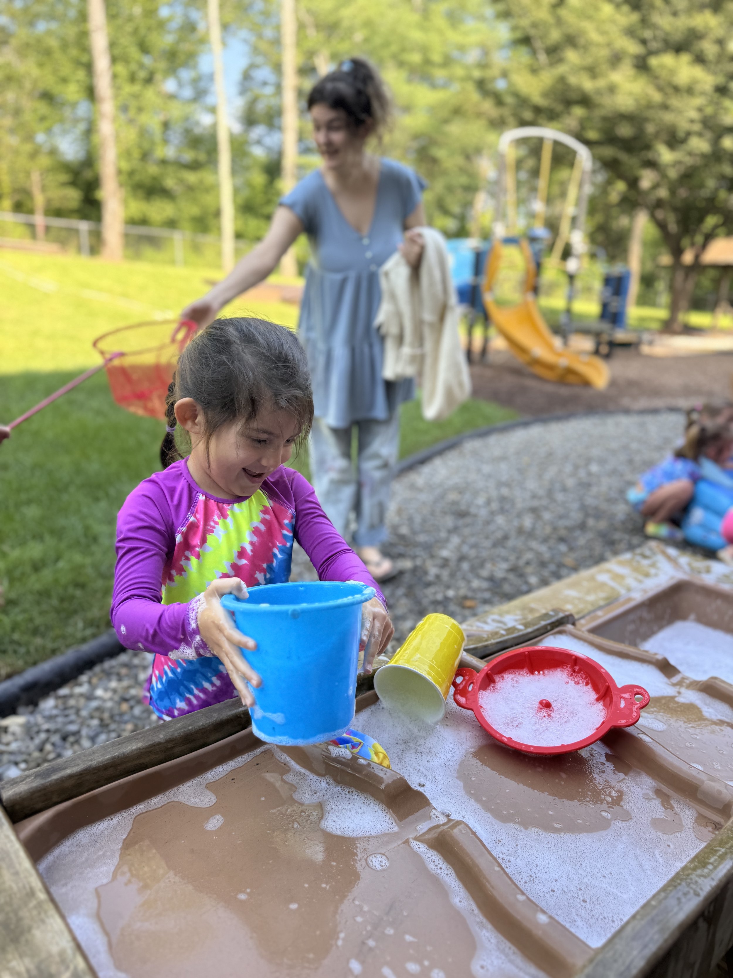 A young girl playing in a water table with colorful buckets and toys, smiling and enjoying outdoor play. A woman is in the background holding a towel and a net, near a playground slide in a park with green trees.