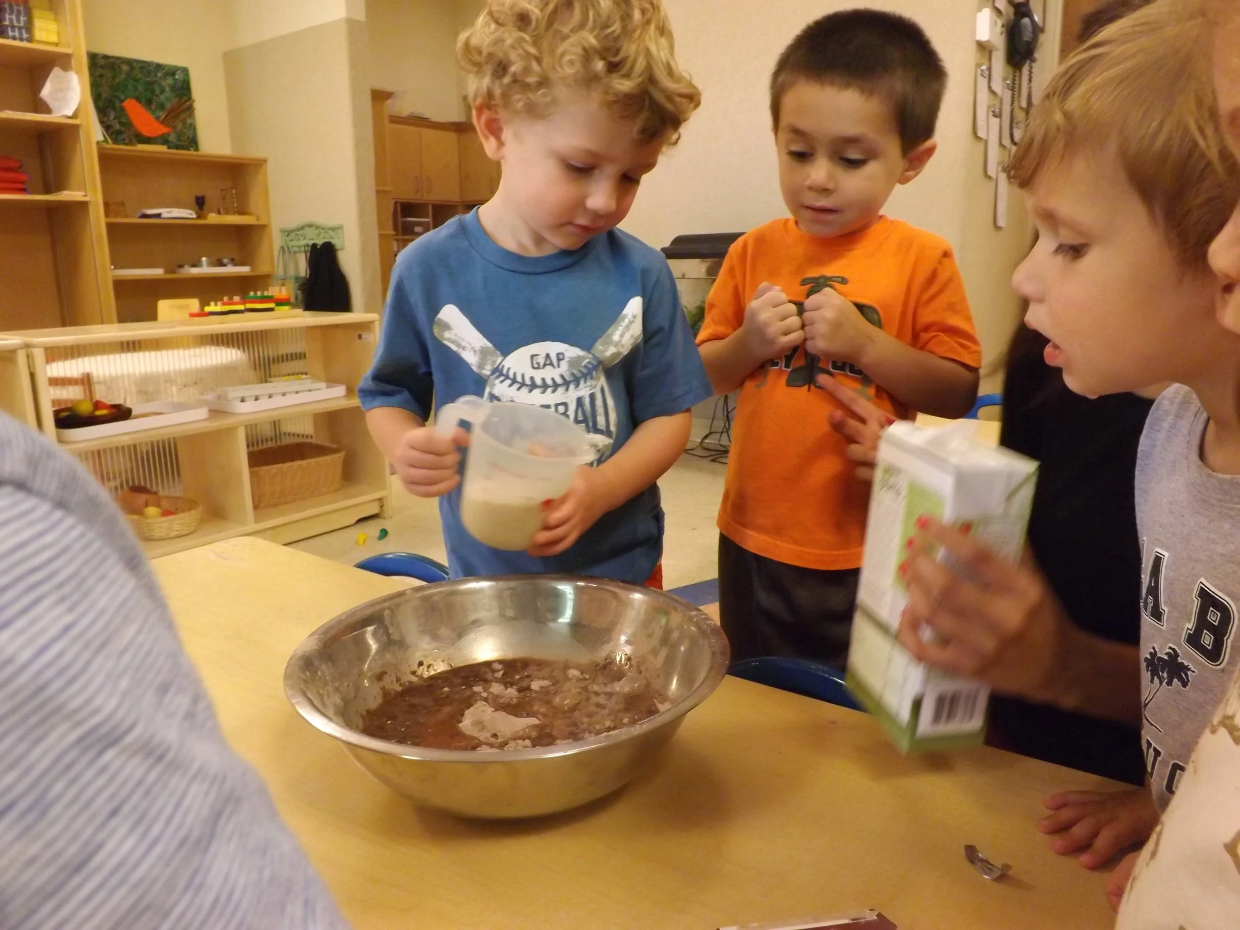 Children gathered around a table, adding ingredients to a mixing bowl for cooking or baking.