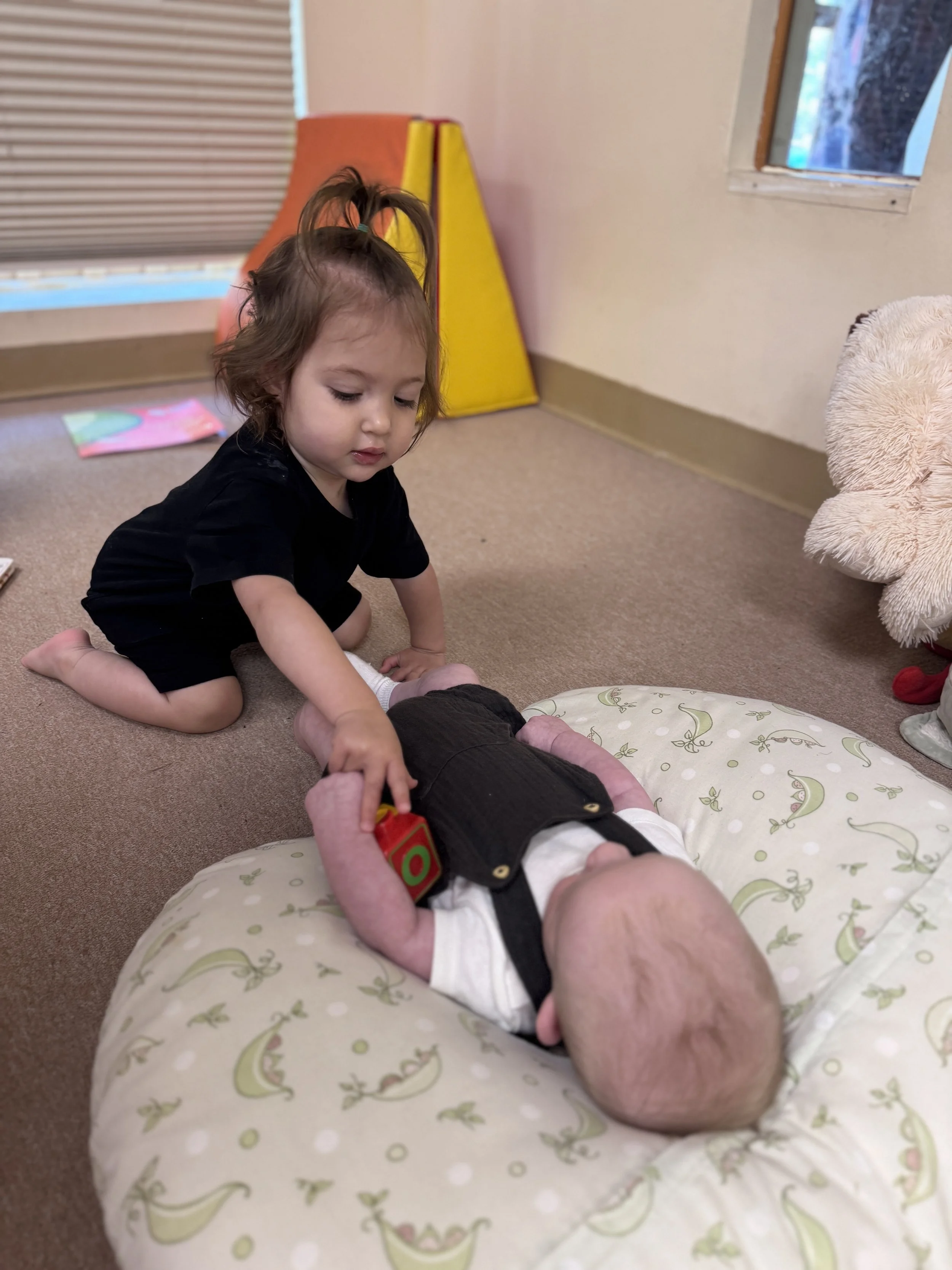 A young girl playing with a baby on a beige carpeted floor in a room with pink walls, a window, and a stuffed animal.