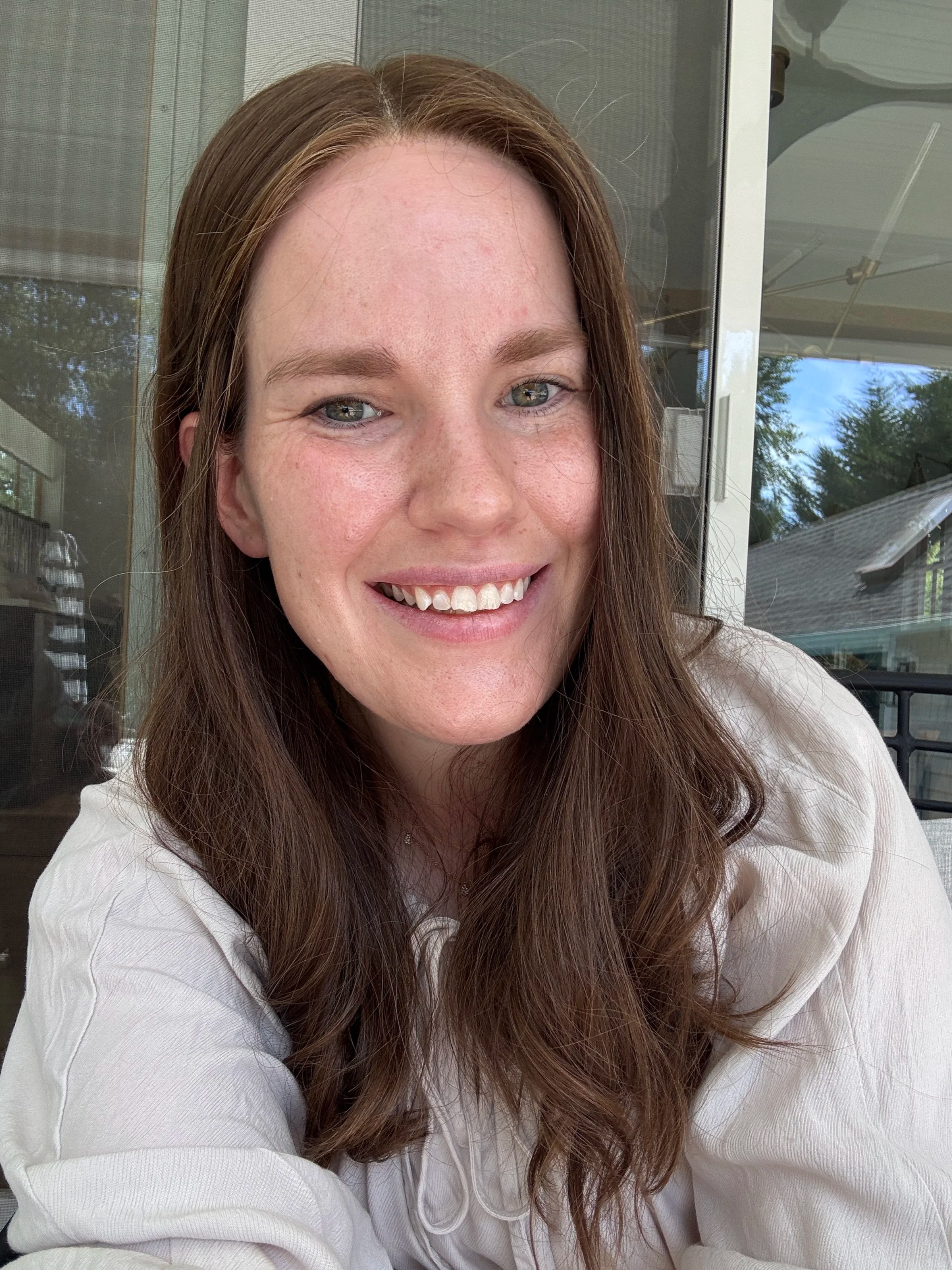 A woman with long brown hair, blue eyes, and freckles smiling outdoors near a glass door and window, with trees and a house visible in the background.