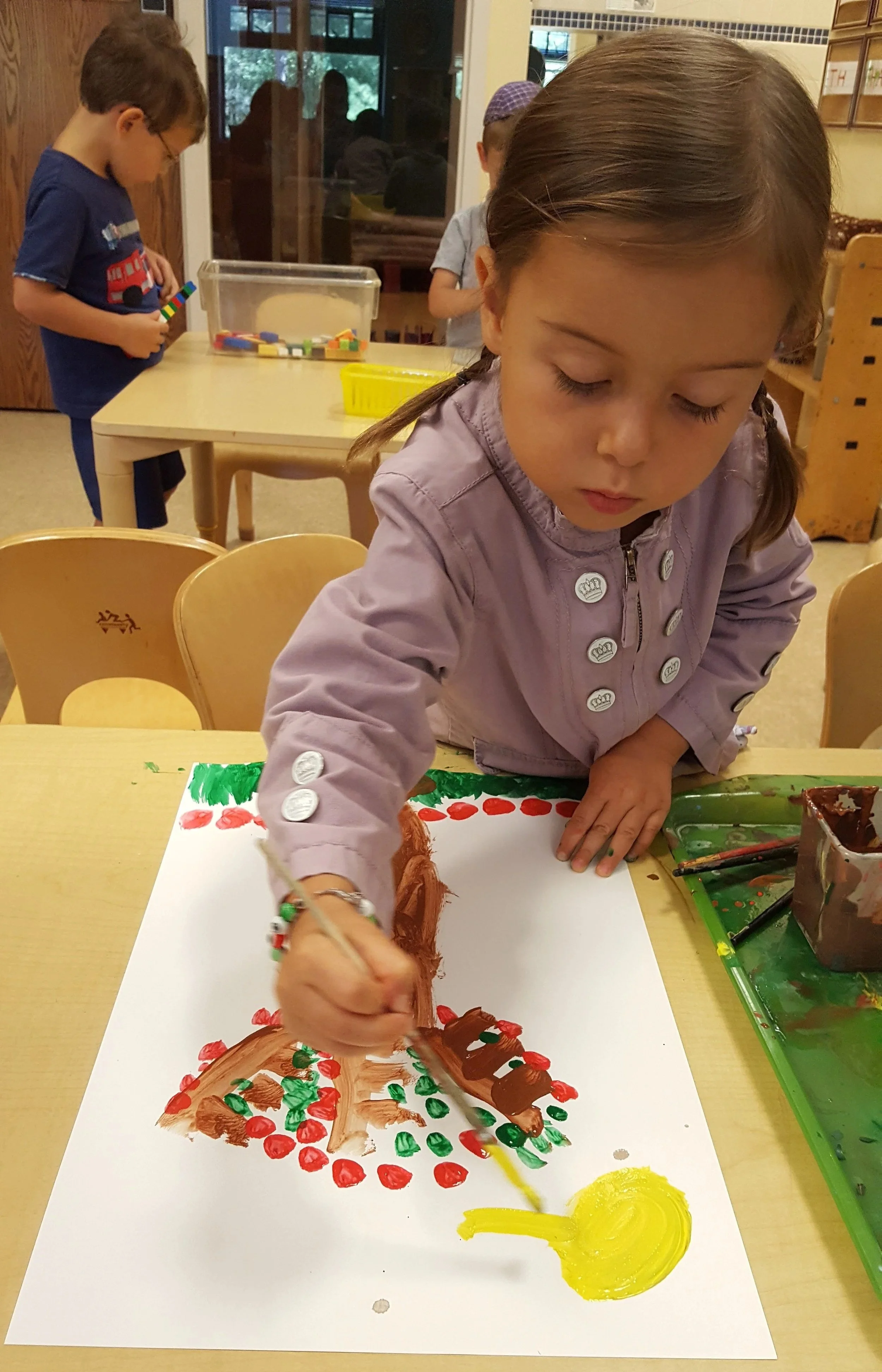 Young girl painting a colorful picture of a Christmas tree with a yellow sun in a classroom.