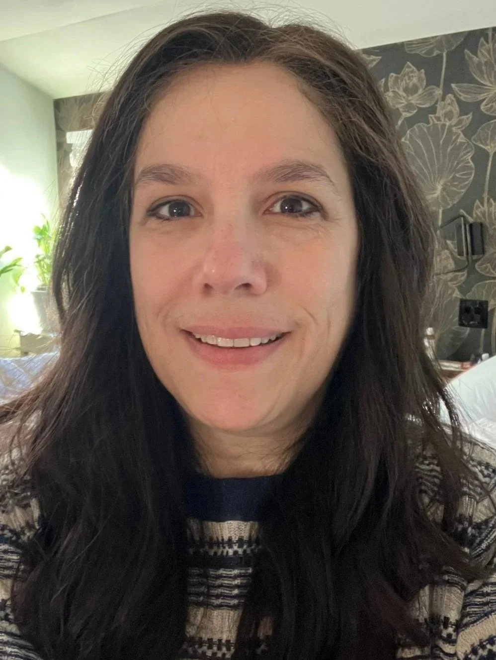 A woman with long, dark curly hair smiling at the camera in a well-lit room with a patterned accent wall and houseplants in the background.