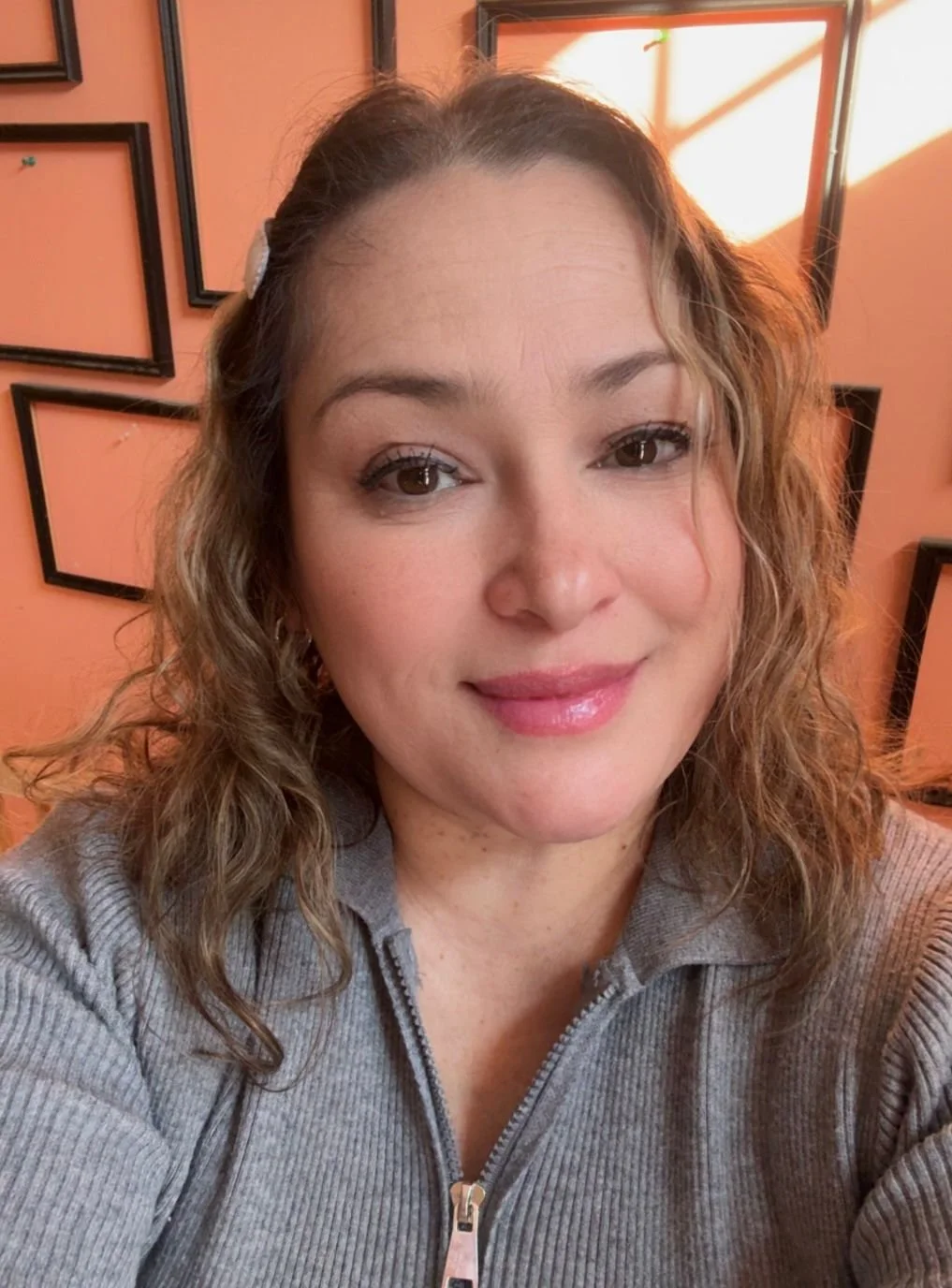 A woman with light skin, curly light brown hair, and brown eyes taking a selfie indoors. She is wearing a gray zip-up sweater and has a soft smile.