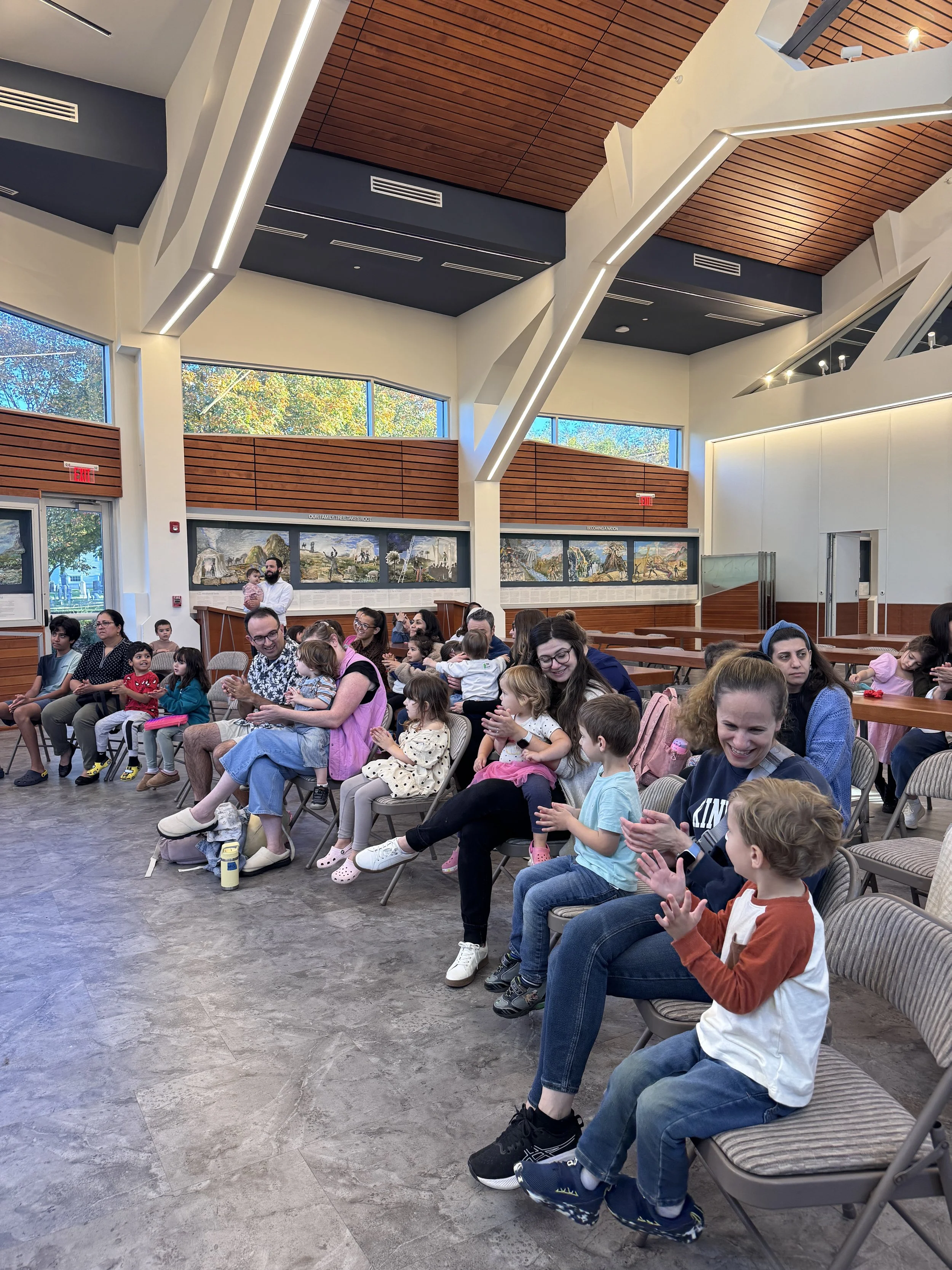 Children and adults sitting in a spacious, modern room with large windows, clapping and smiling during an indoor event.