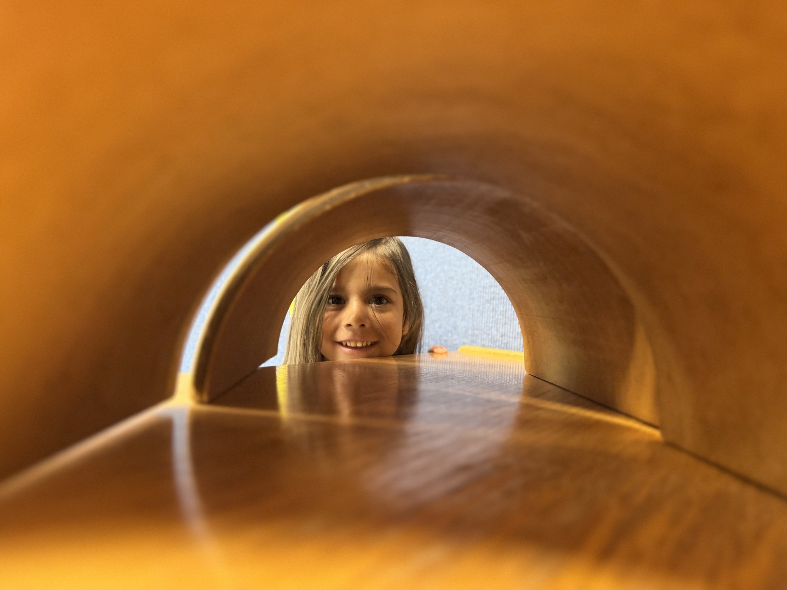 Young girl with long hair smiling, visible through the arch of a wooden tunnel or tube, with a blurred background.