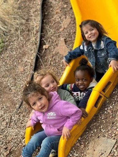 Four young girls sitting on a yellow playground slide outdoors on dirt and rocks, smiling.