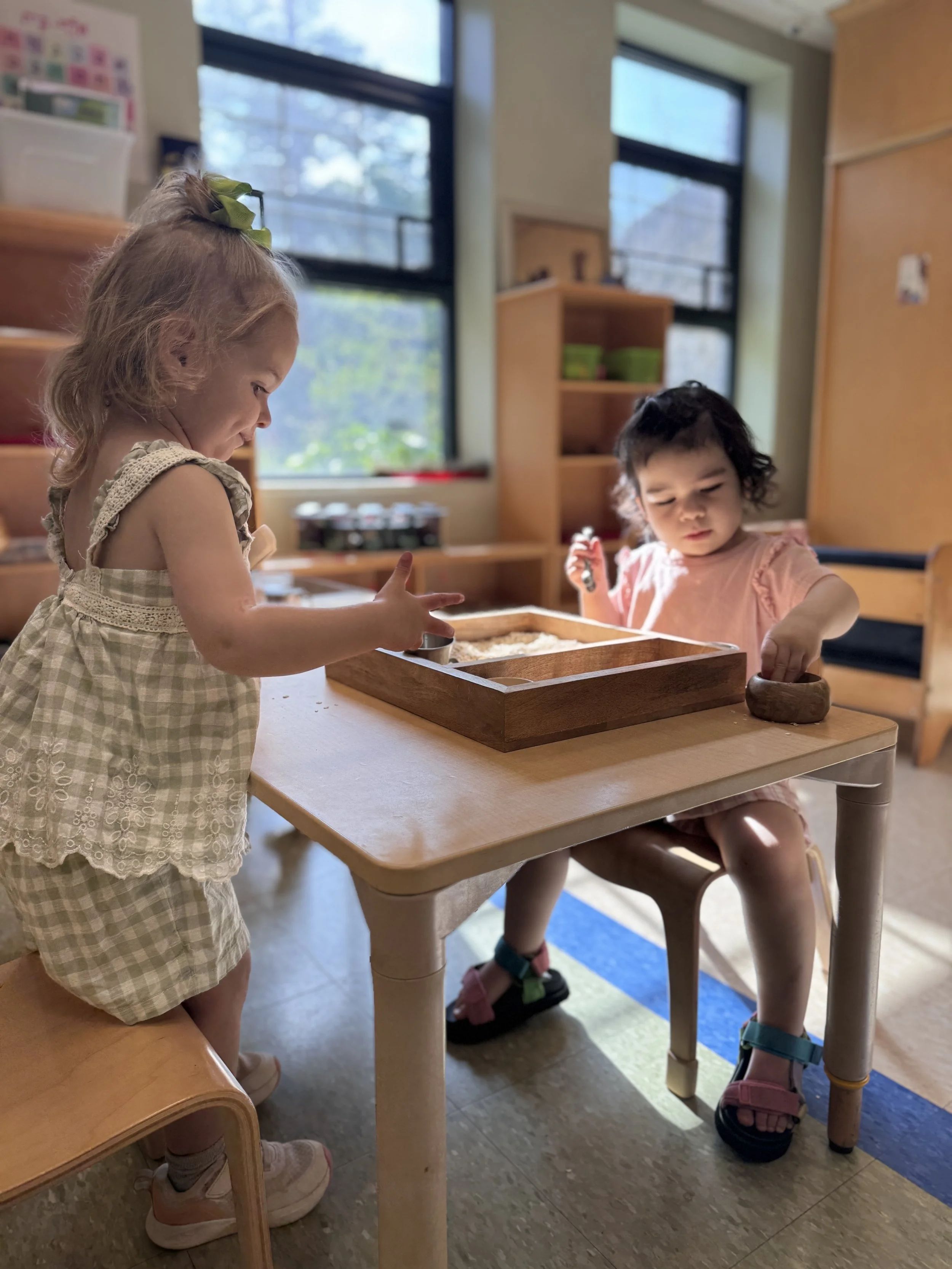 Two young girls playing with toys at a table in a classroom with large windows and wooden furniture.