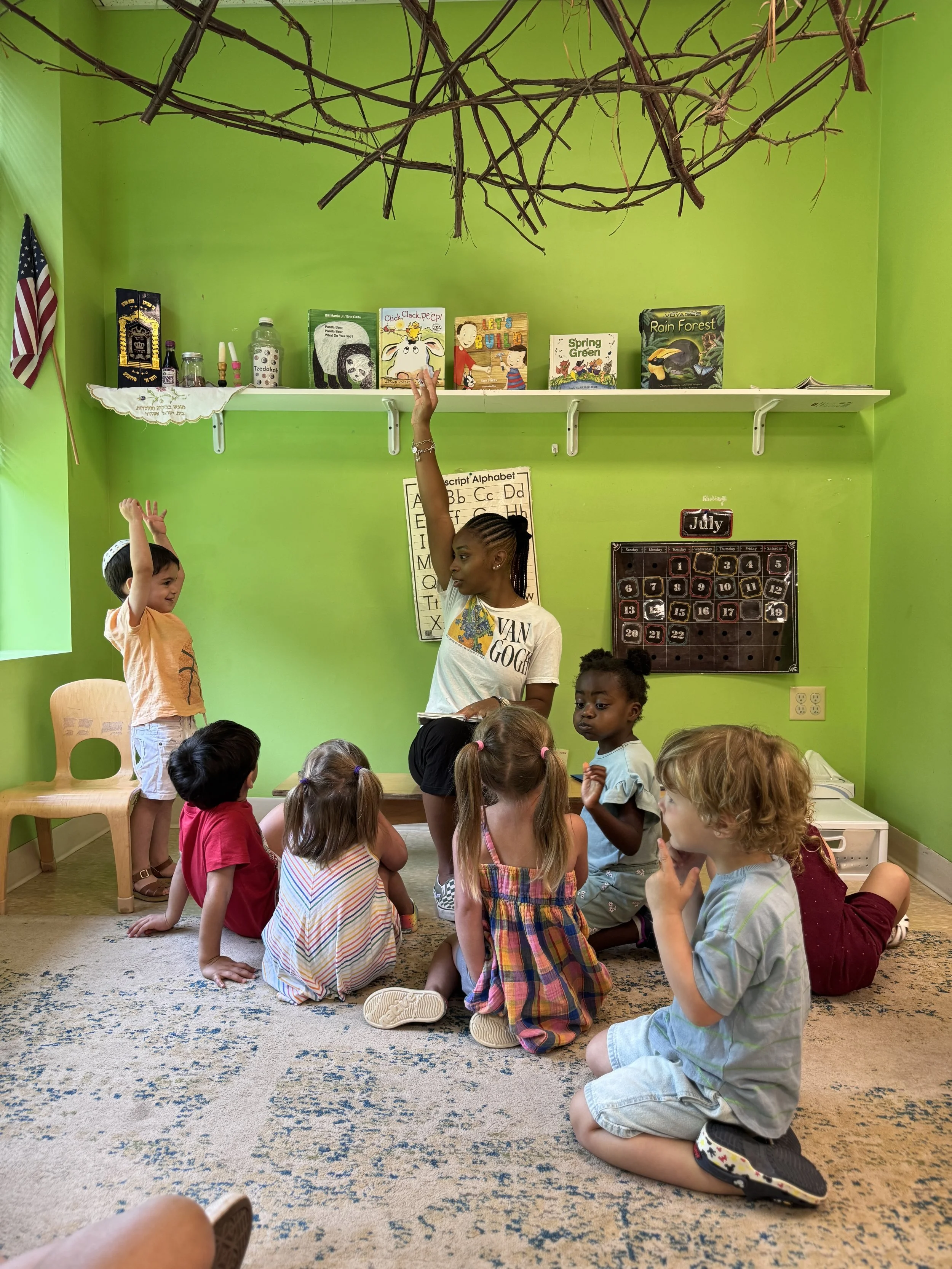 A classroom with a green wall, a teacher engaging children sitting and kneeling on the carpet. The teacher is reaching to a shelf with books, and children are participating in an activity.