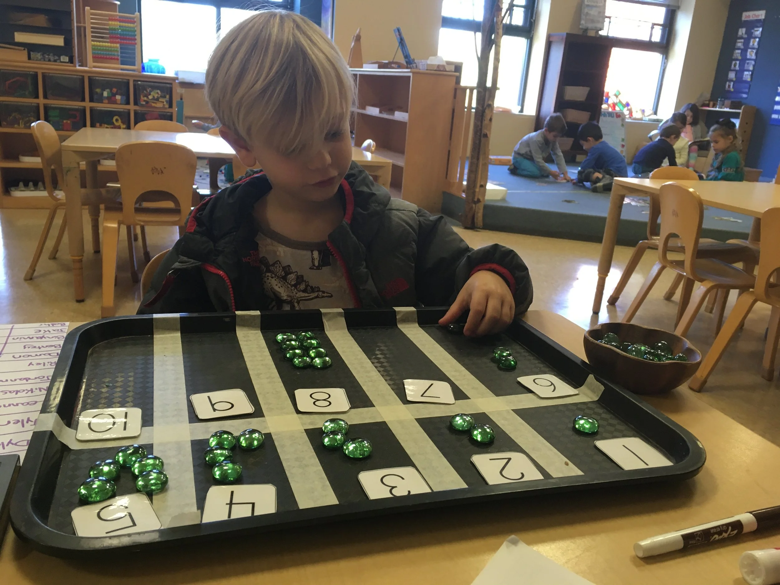 A young boy in a black jacket is playing a game with green glass stones and numbered cards on a black tray in a classroom. In the background, several children are sitting on a mat, engaged in an activity.