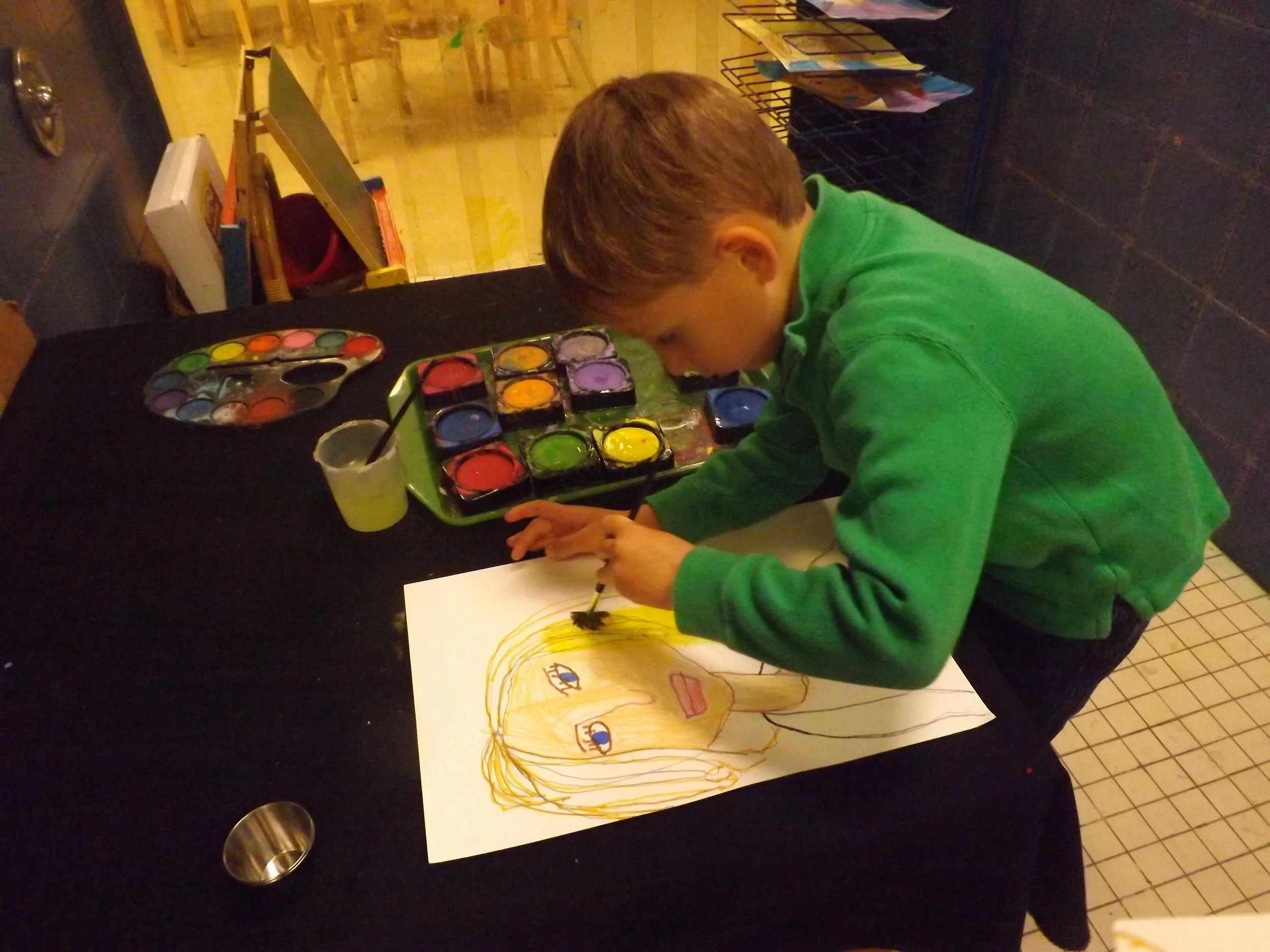 A young boy in a green sweatshirt is painting a colorful portrait on a piece of paper, using watercolor paints on a black table.