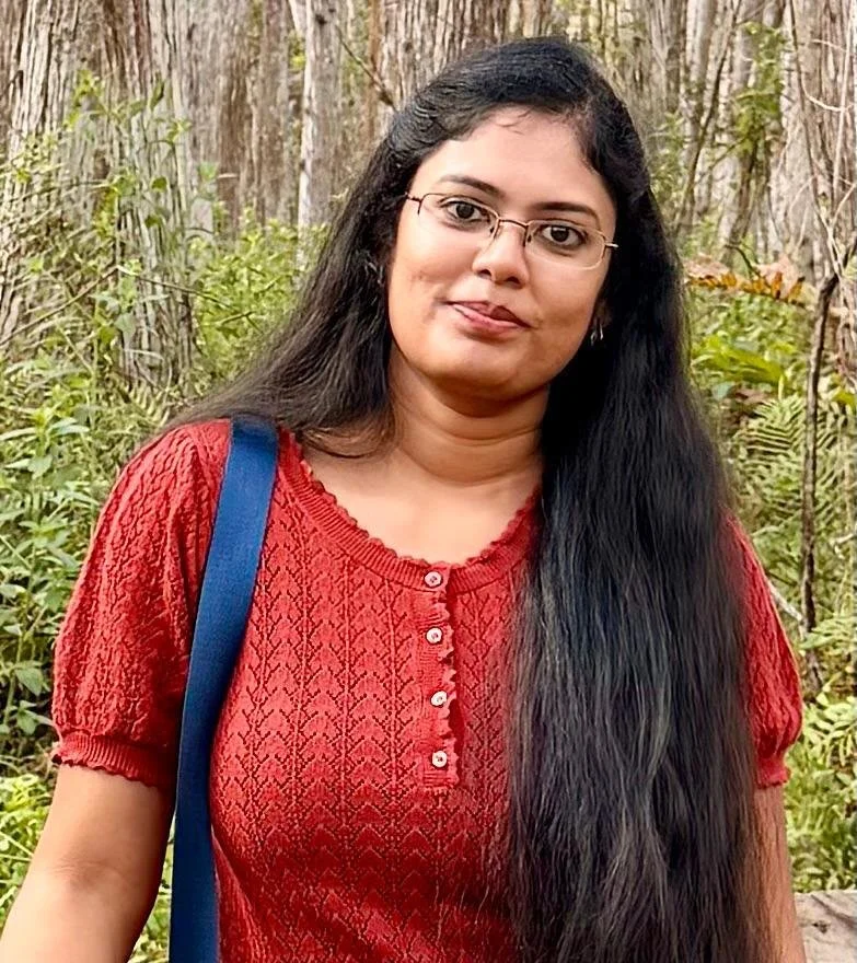 A woman with long black hair, glasses, and a red textured top standing outdoors in a wooded area with green foliage.