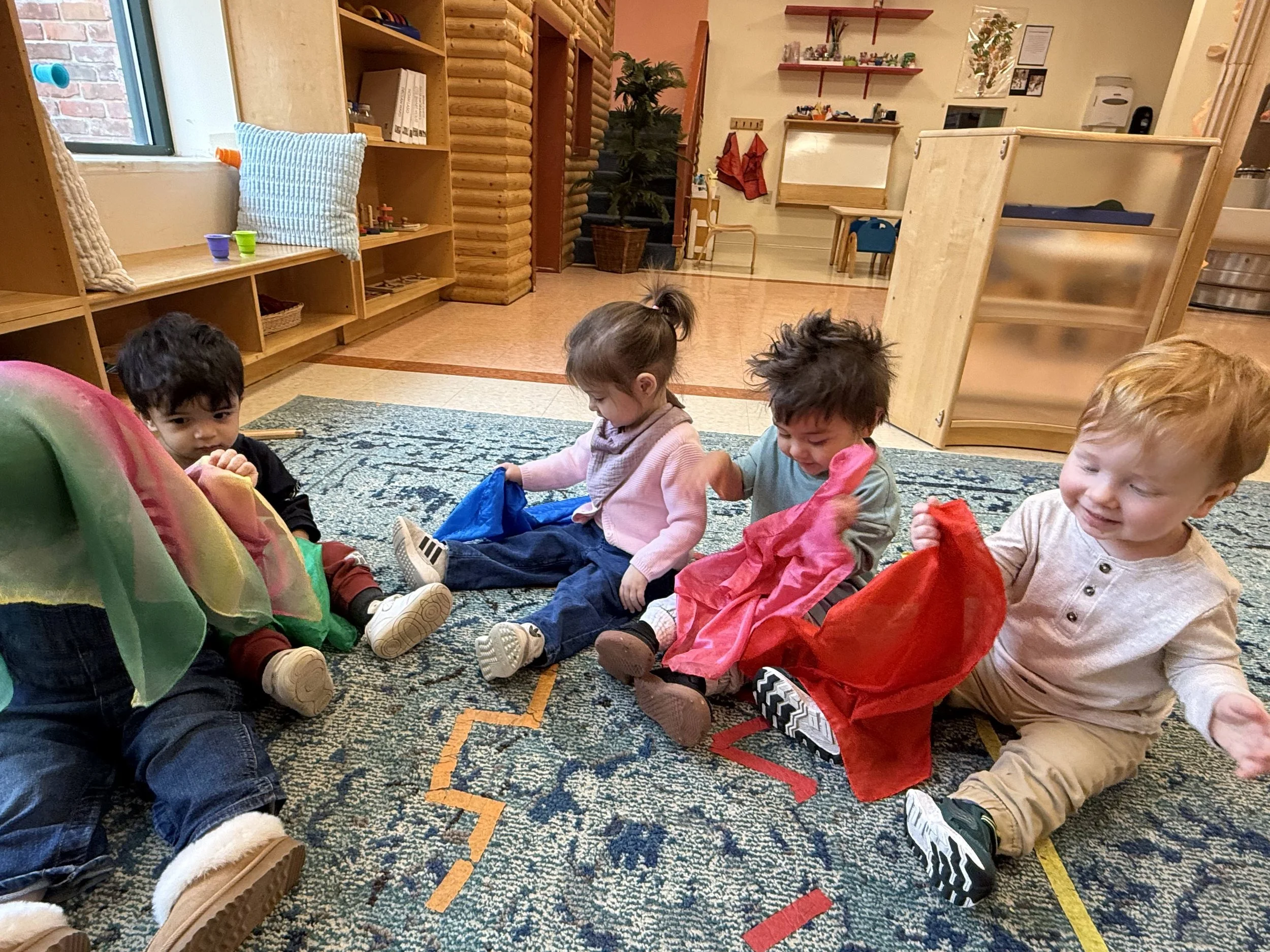 Four young children sitting on a colorful rug in a classroom, holding various colored cloths, with shelves and educational materials in the background.