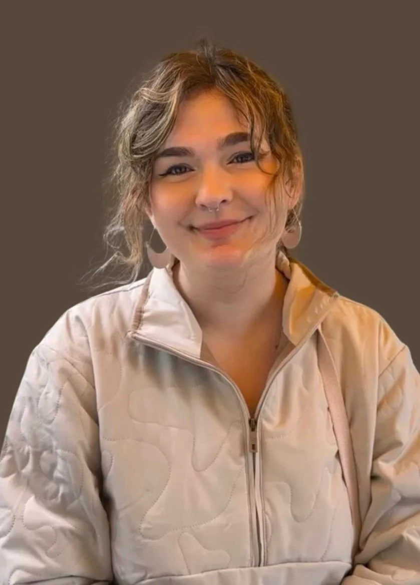 A woman with wavy, light brown hair, wearing a beige quilted jacket, smiling with a neutral brown background.
