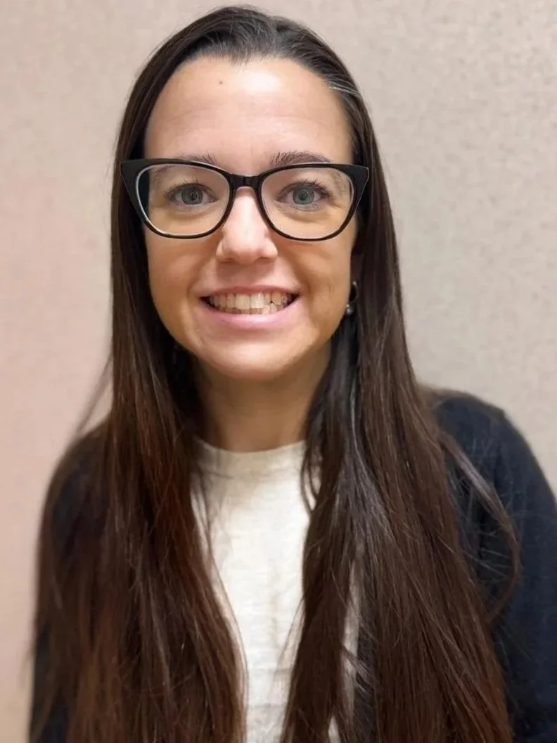 A woman with long brown hair, wearing glasses, smiling, and dressed in a light-colored top with a dark sweater, standing against a beige wall.