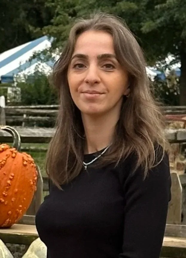 A woman with brown hair, wearing a beige turtleneck sweater, smiling at the camera, background with green and blue walls.